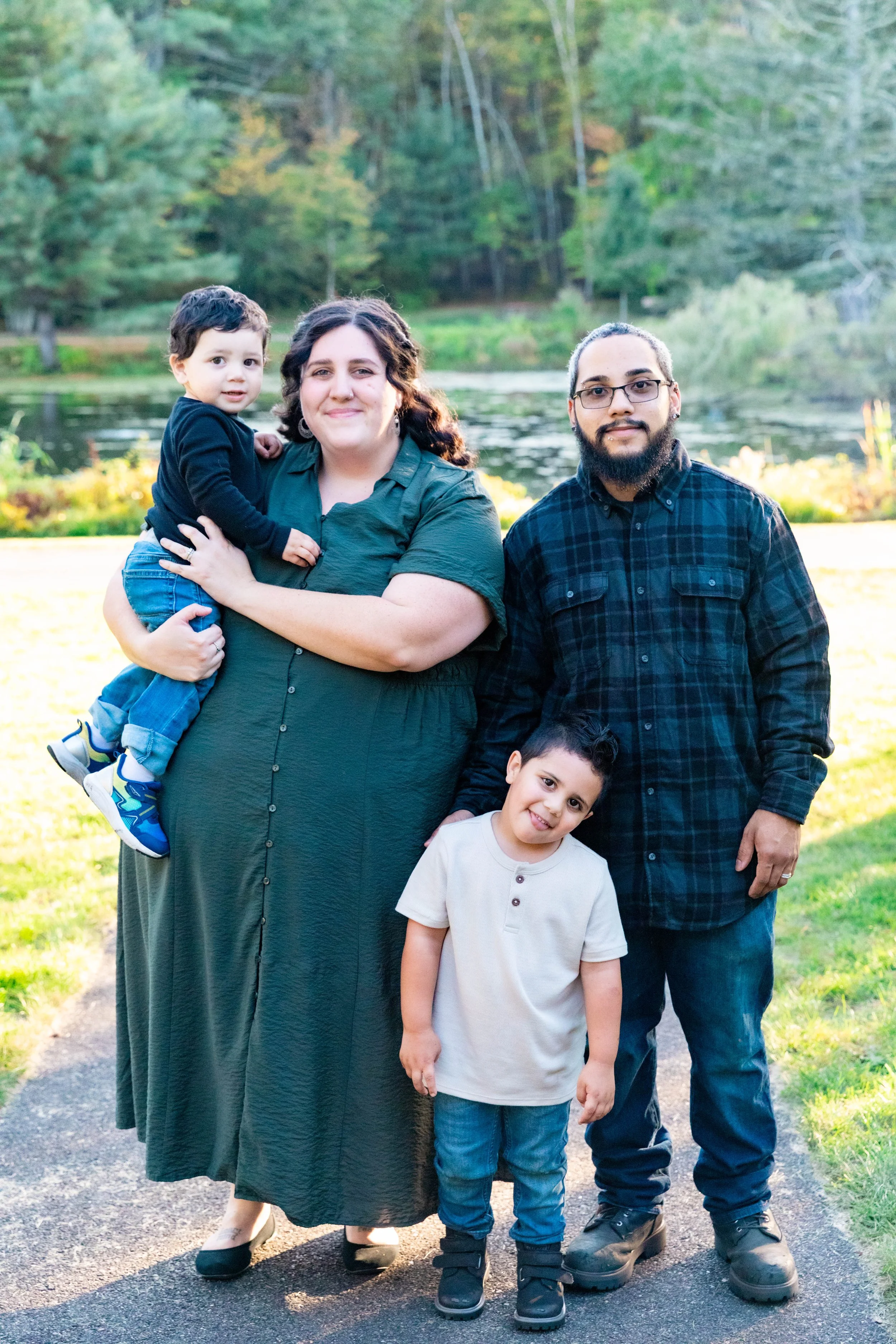 A family of four posing outdoors near a pond, with trees in the background during daytime.