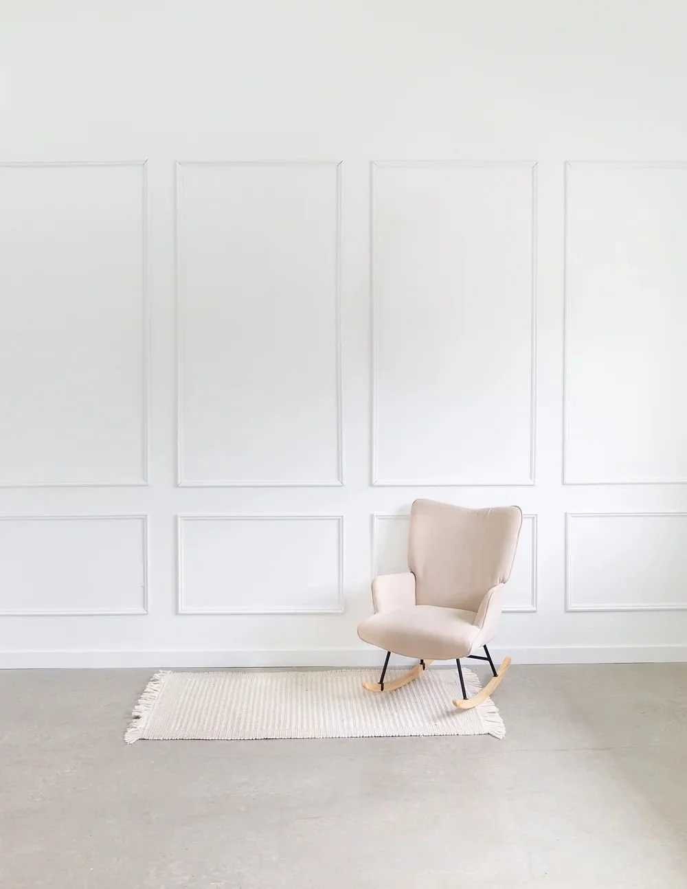 A beige rocking chair on a small cream rug against a white paneled wall.