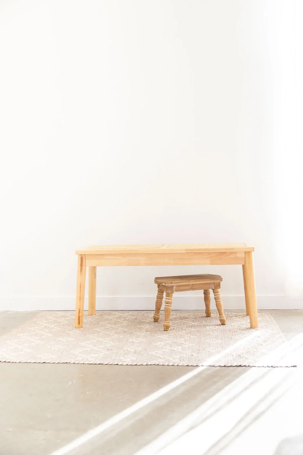 A light wood table and a small matching stool placed on a beige rug in front of a plain white wall, with sunlight casting shadows on the floor.