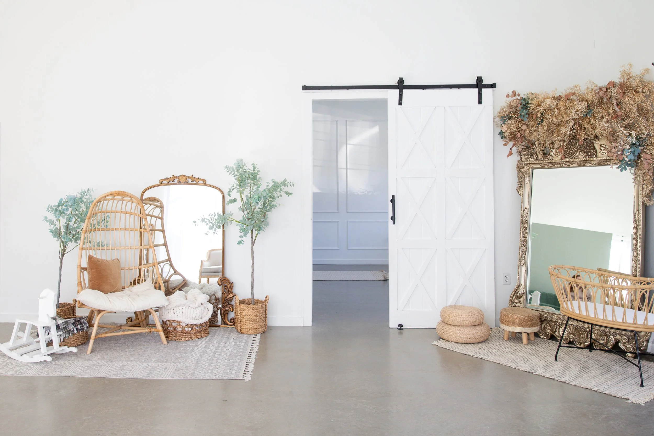 Decorative room with wicker furniture, plants, a mirror, a large mirror with an ornate gold frame, and dried flowers, all arranged on area rugs against white walls.