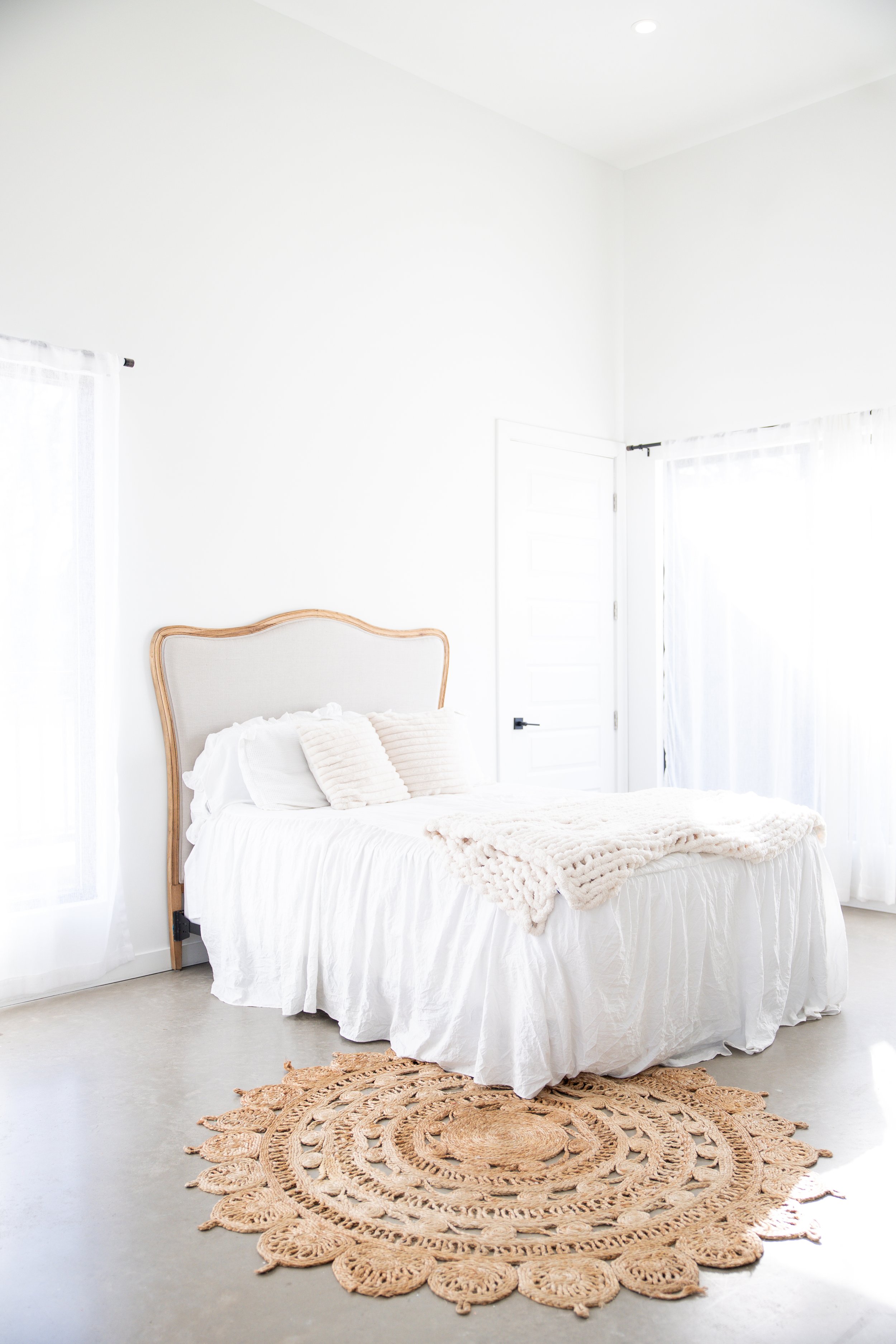 Bright white bedroom with a bed covered in white linens and a beige headboard. There are two white pillows and a beige blanket on the bed. A beige circular rug with intricate patterns is on the floor. The room has sheer white curtains on both windows