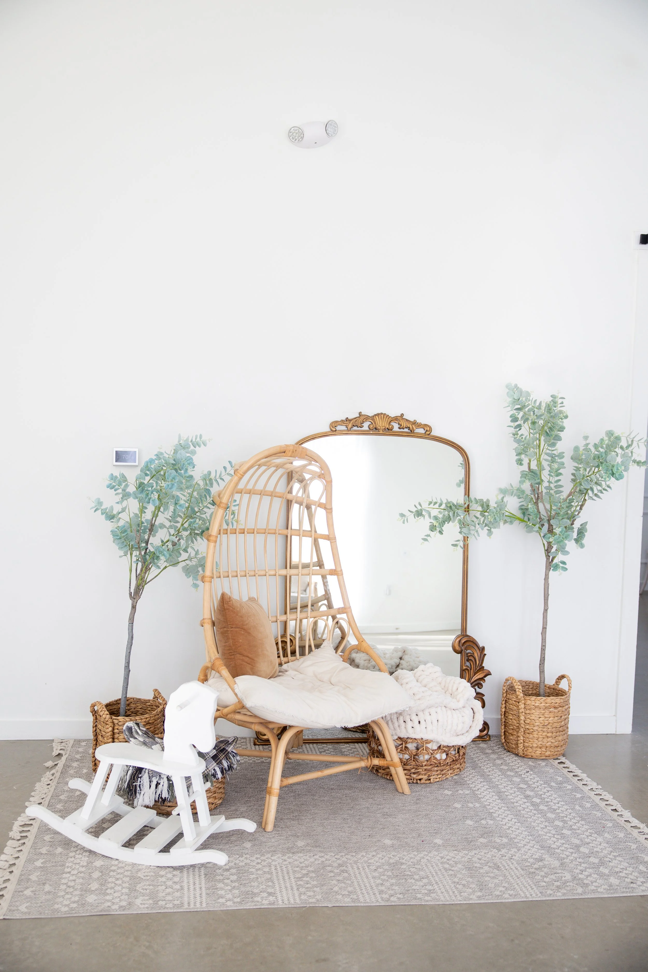 Decorative interior corner with two potted faux eucalyptus trees flanking a woven rattan chair with a pillow and cushion, a white white rocking horse, a large ornate mirror, and a woven basket on a patterned rug, against a plain white wall.
