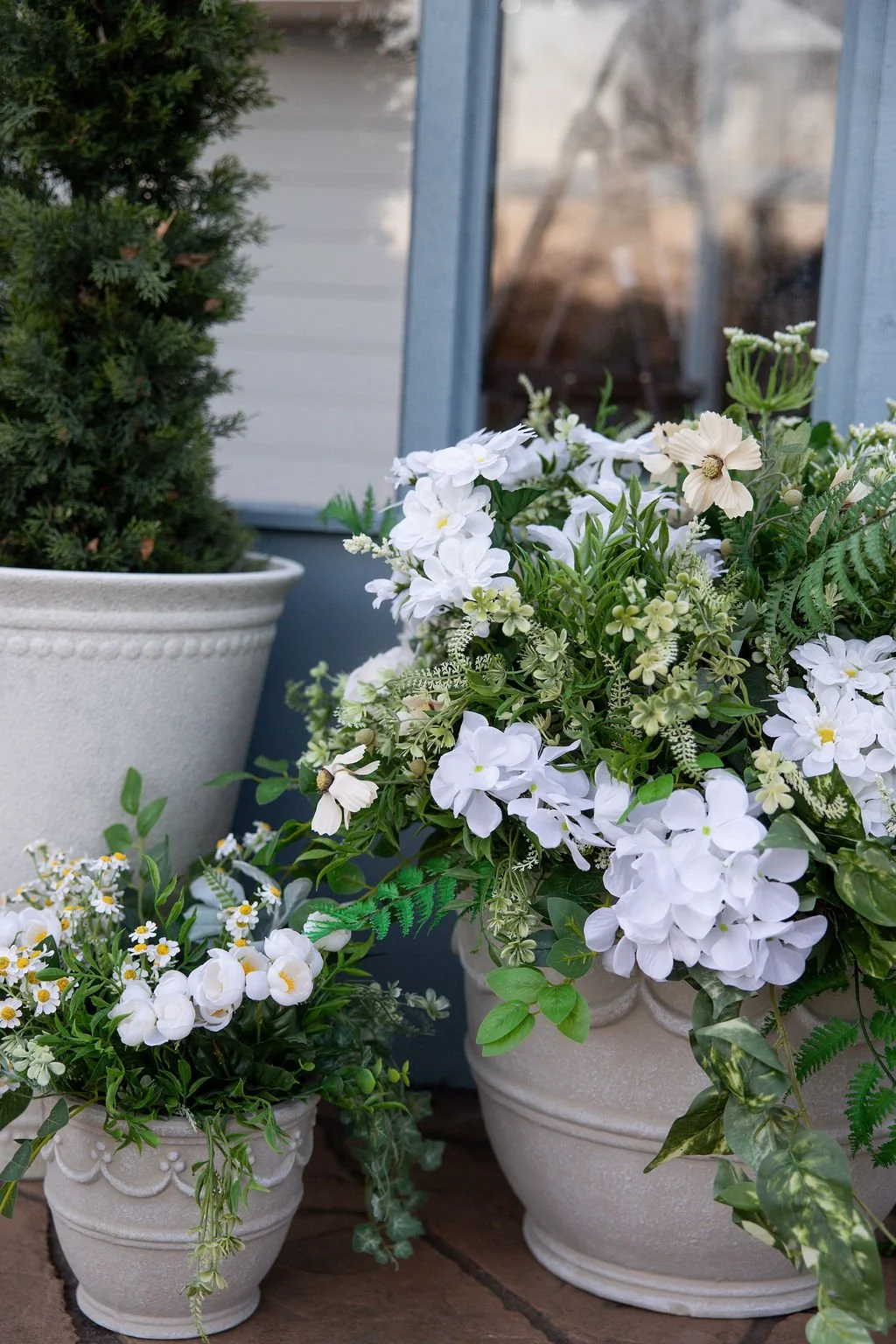 Potted white flowers and greenery outside a house with a window in the background.