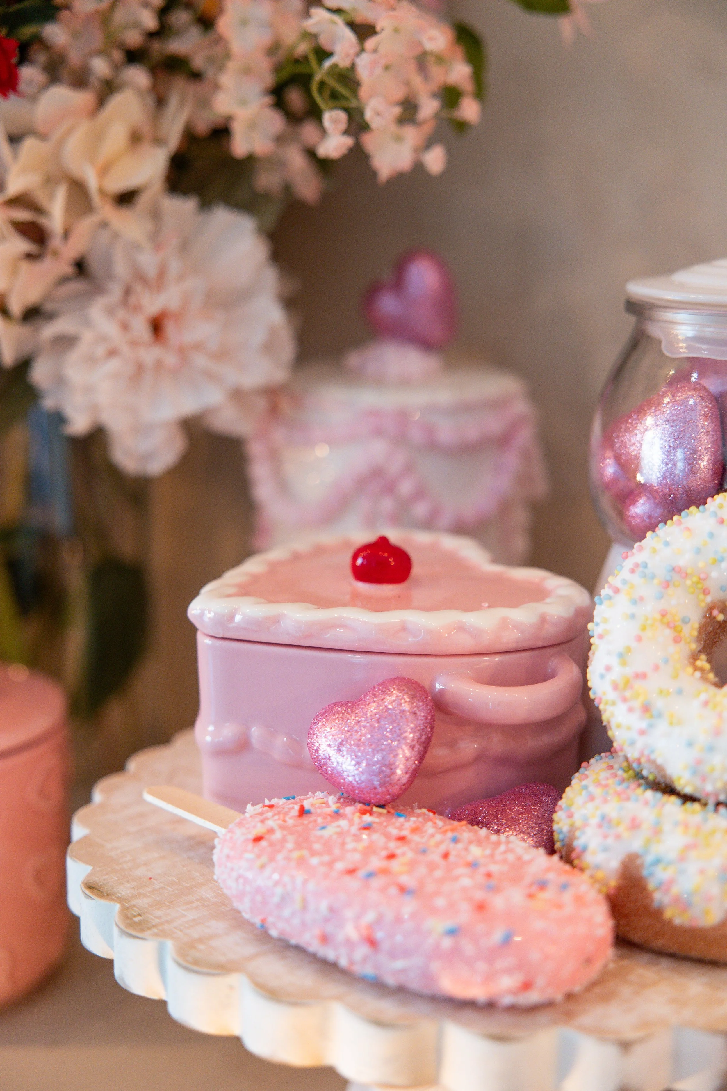 Various pink and white Valentine's themed treats, including a pink mug-shaped container with a cherry on top, decorated cookies with pink and heart-shaped sprinkles, a jar filled with pink glittered heart-shaped candies, and a pink frosted cake with 