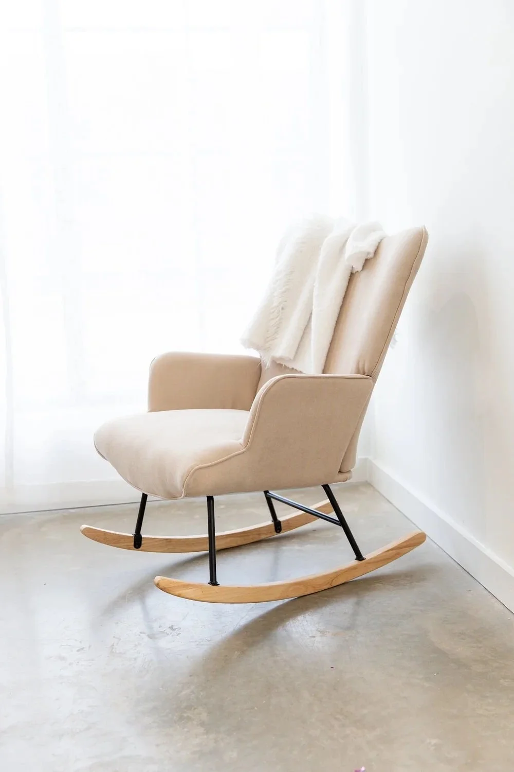 Beige rocking chair placed next to a white wall and sheer curtain, with a soft white blanket draped over its backrest.
