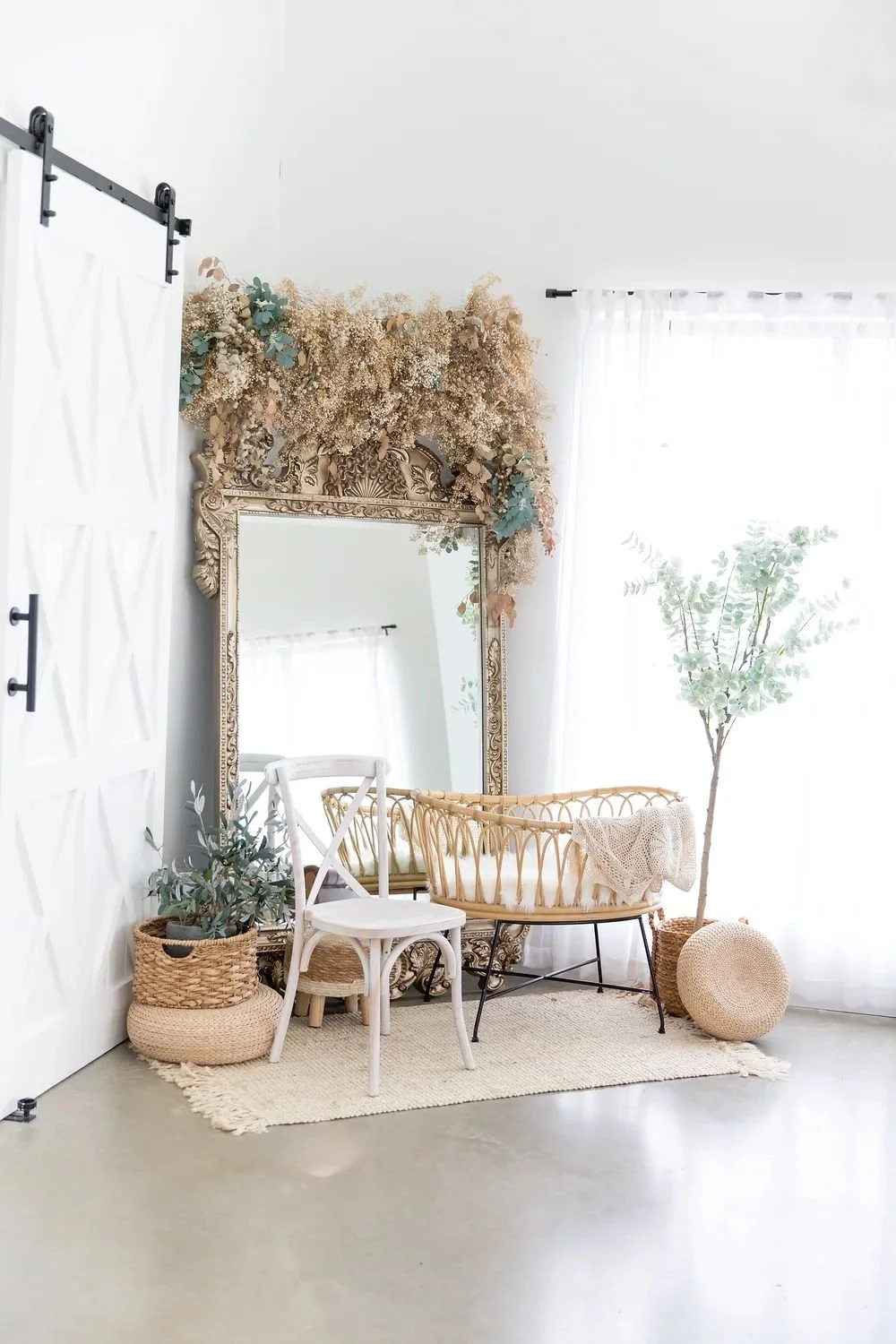 A boho-style decorated room with a large ornate mirror, dried flowers, a white chair, a wicker bassinet with a blanket, potted plants, and sheer curtains letting in natural light.
