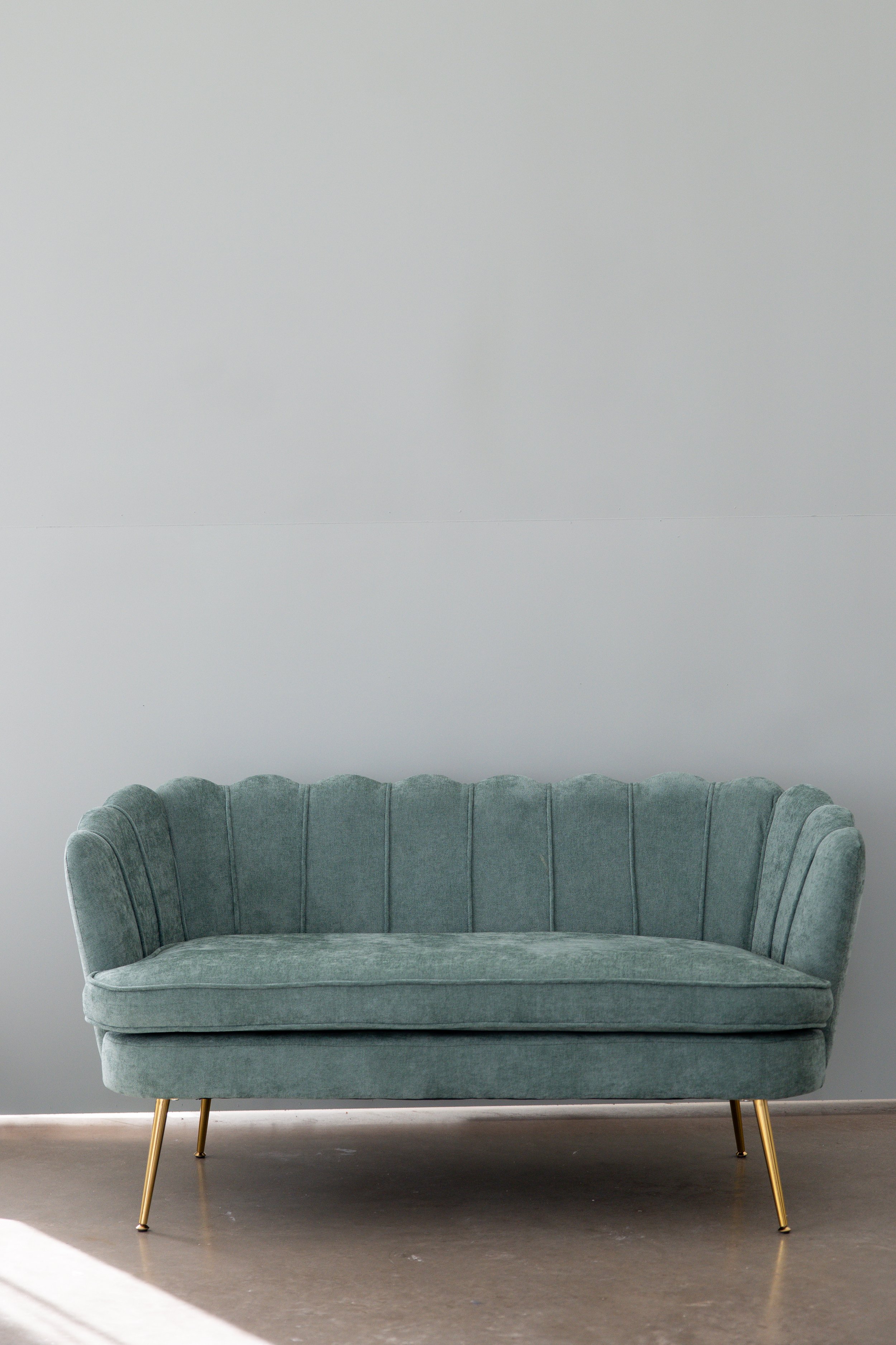 A vintage-style green velvet sofa with gold legs, positioned against a plain white wall on a gray floor.
