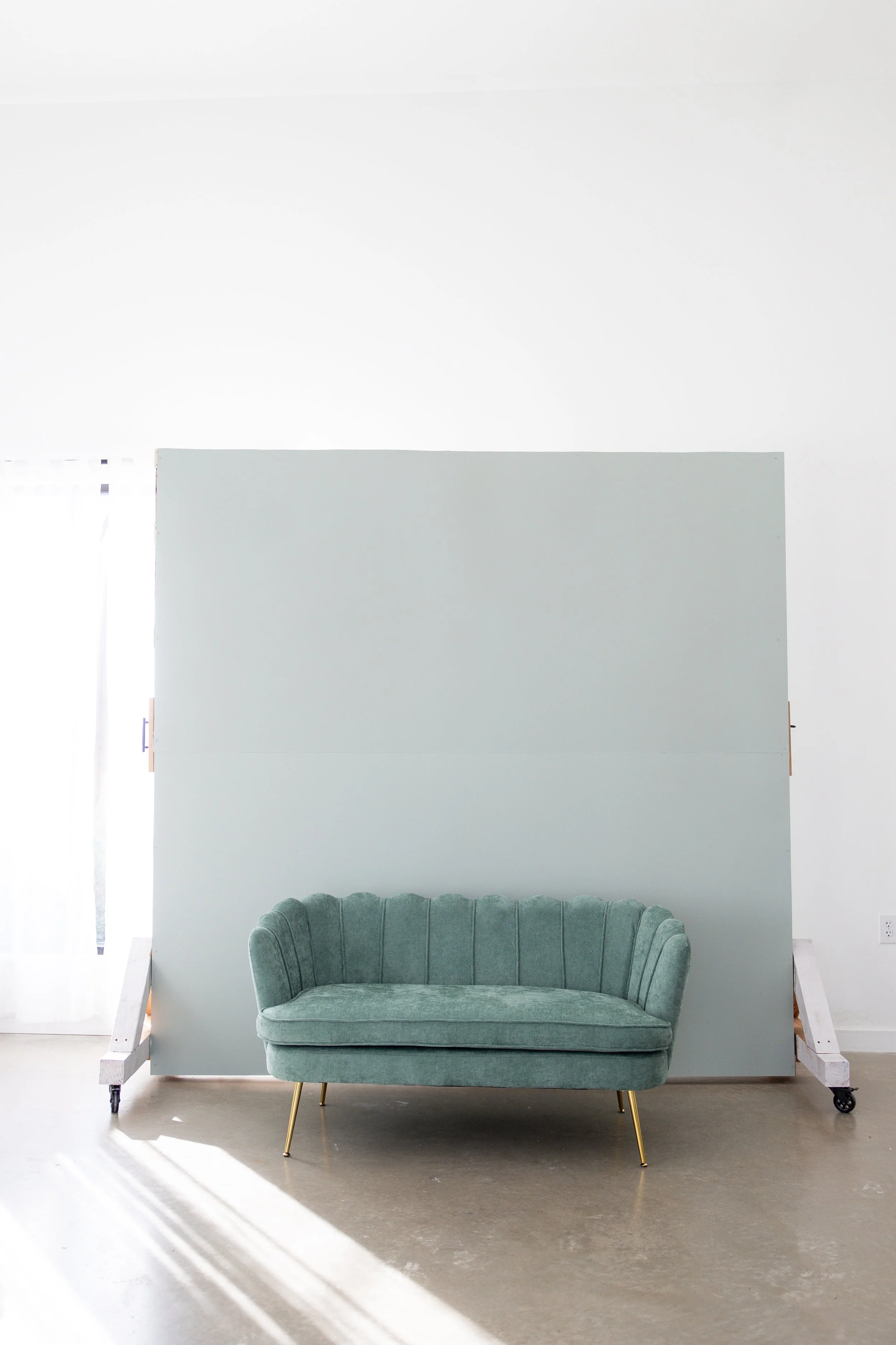 A mint green velvet vintage sofa with gold tapered legs in front of a white partition in a bright room with concrete floor.