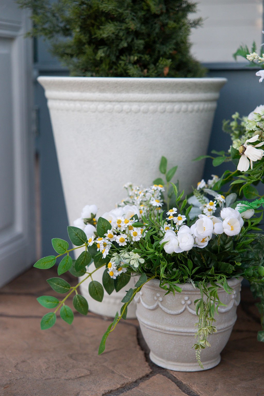 White flowers in a decorative white ceramic pot on a wooden surface, with a larger planter with greenery behind them.