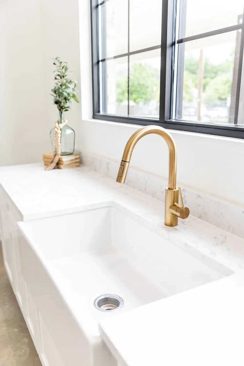 A kitchen sink with a gold faucet, white marble countertop, a window with black frames, and a vase with greenery and beads on the left side.