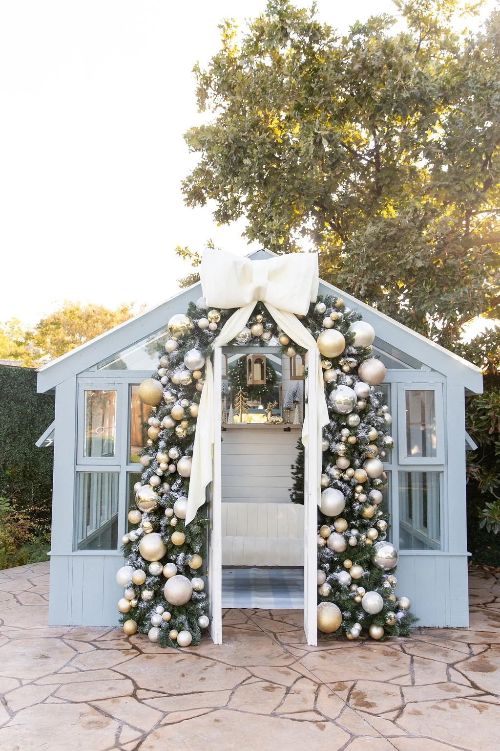 A small light blue wooden greenhouse decorated with white and green floral arrangements, including an arch of flowers above the entrance and potted greenery on either side, placed on a stone patio.
