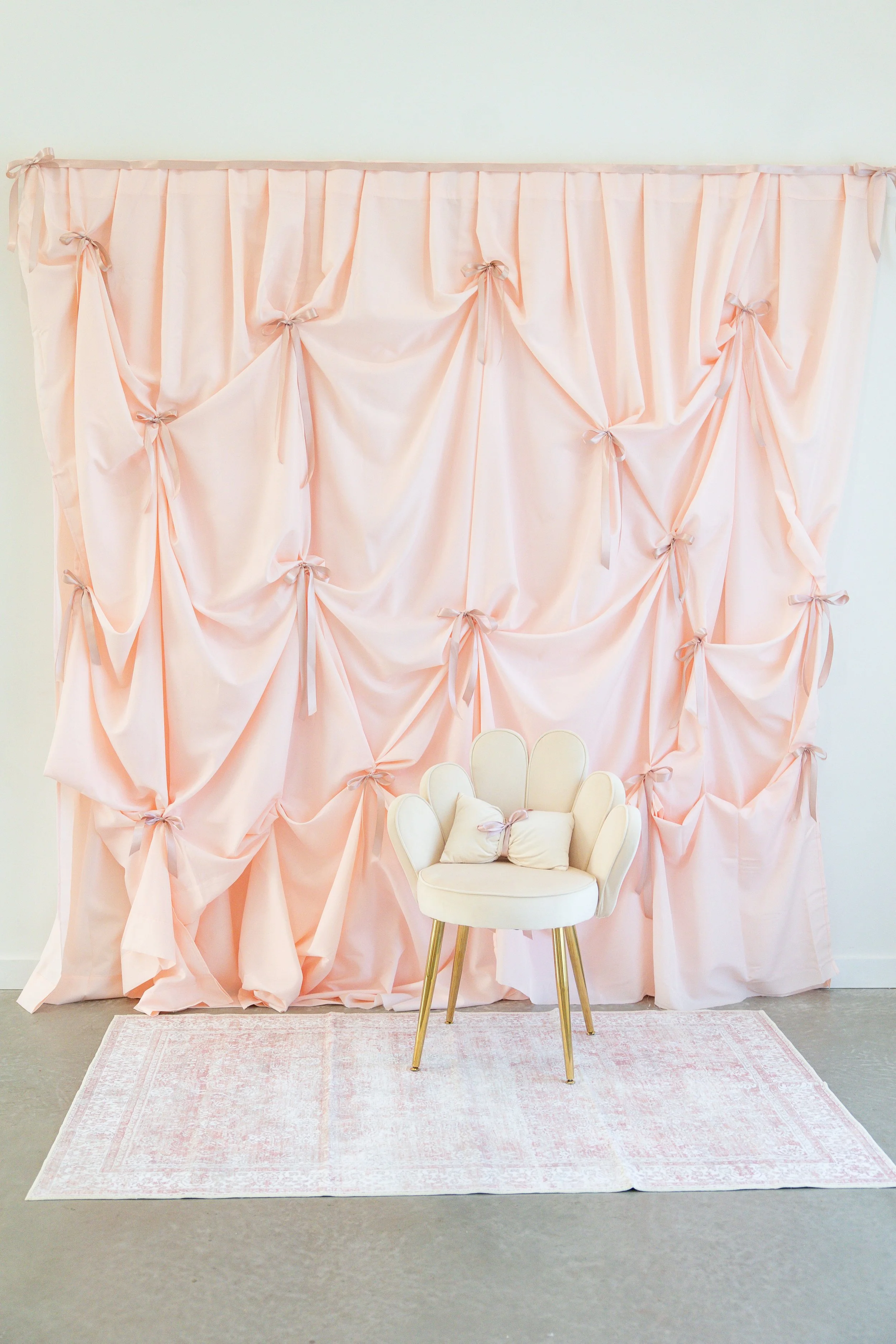 A soft pink, gathered fabric backdrop with bows, set against a plain wall. In front of the backdrop, there's a small, vintage-style cream-colored chair with gold legs, adorned with two small pillows, placed on a light pink rug.