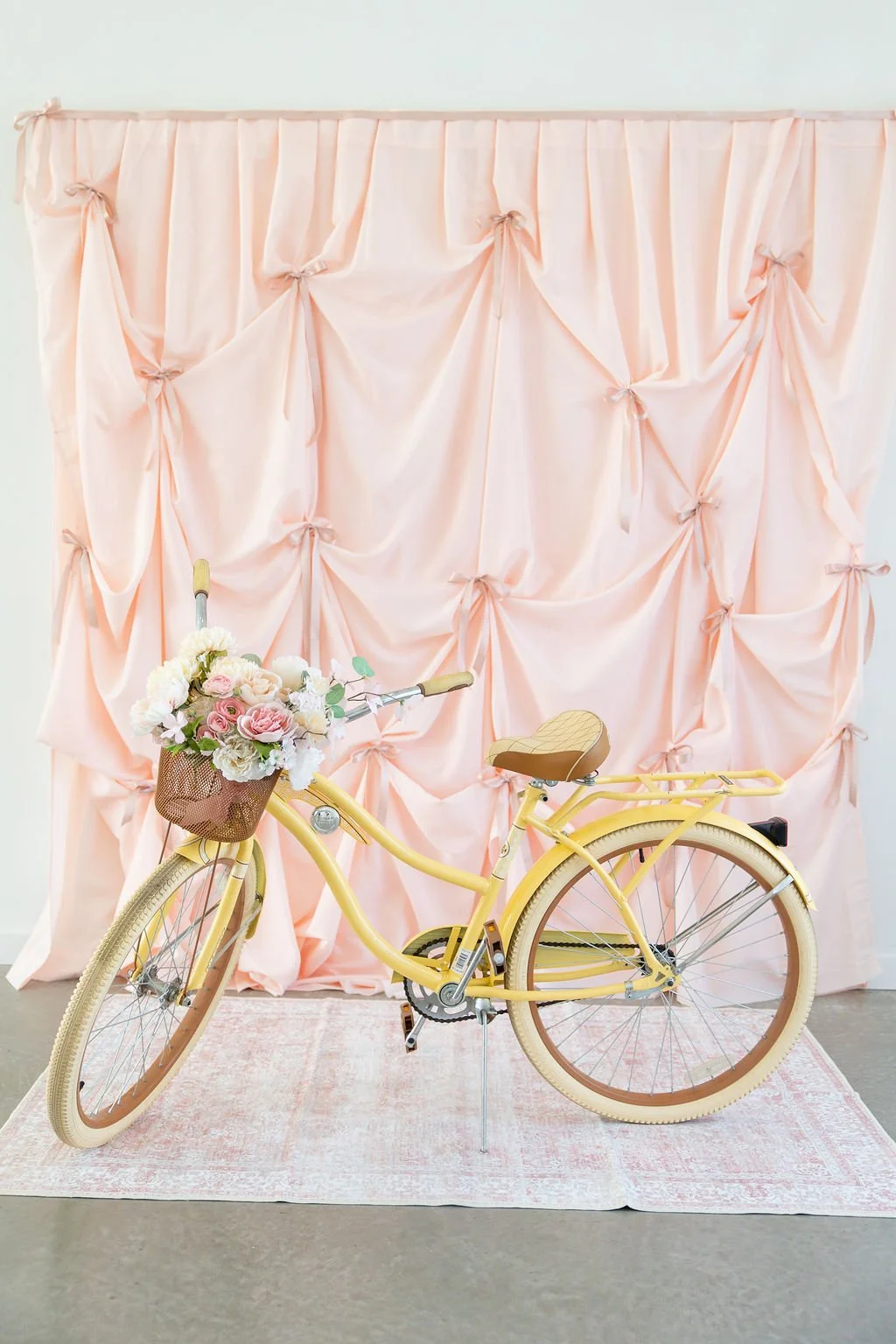 Yellow bicycle with a flower basket in front of a pink draped fabric backdrop.