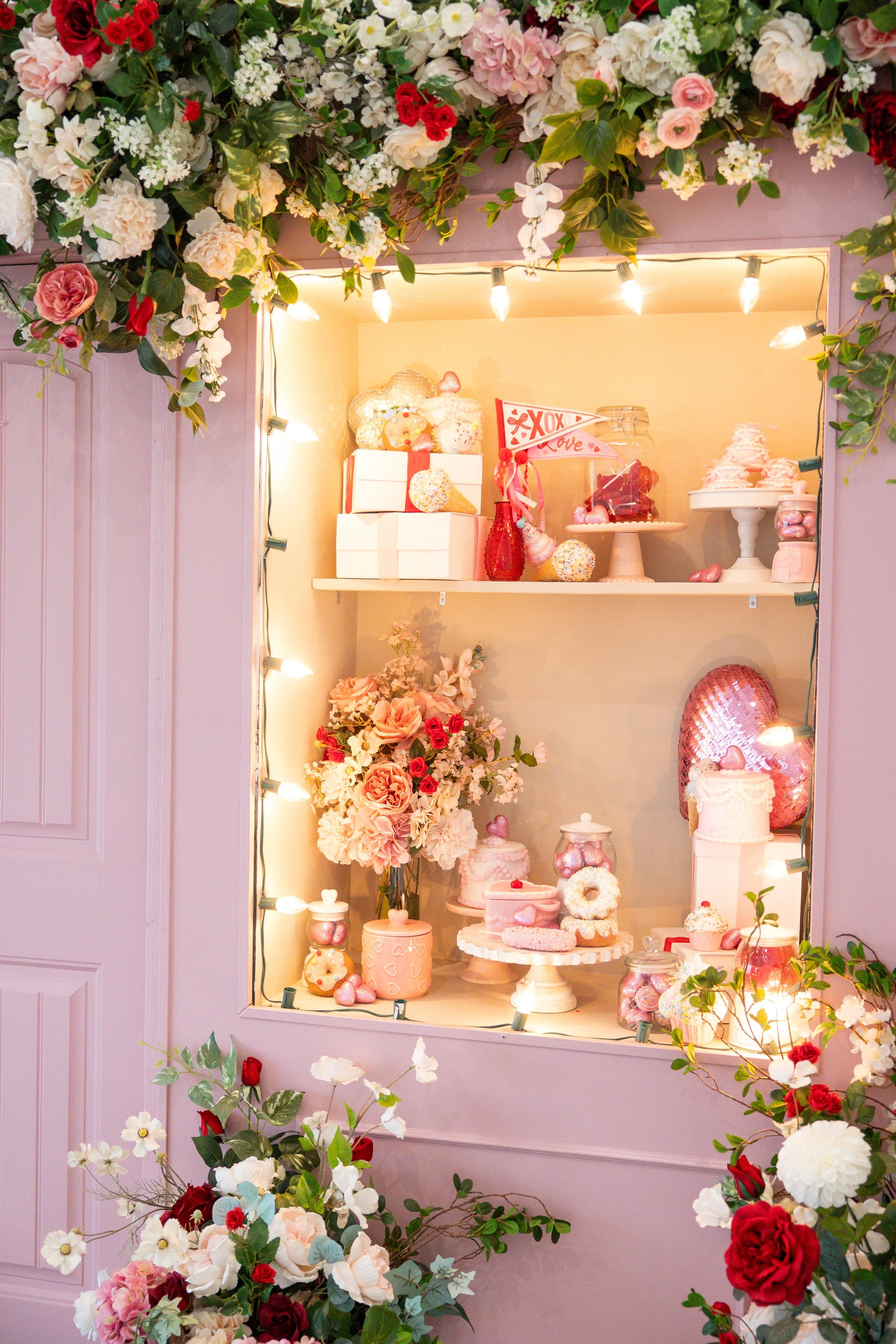 Display of pink, red, and white flowers with Valentine's Day-themed desserts and decorations, illuminated by string lights.