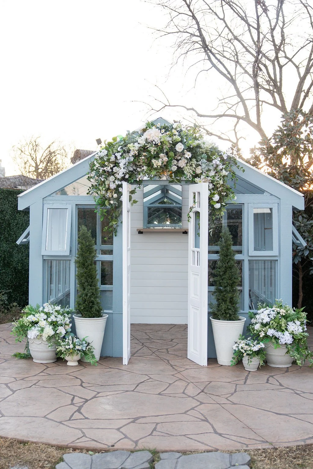 A small light blue greenhouse decorated with white flowers and greenery, with open white double doors and potted plants on a stone patio, during daytime.