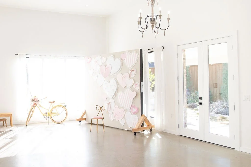 Bright, minimalistic living room with white walls and flooring, a chandelier hanging from the ceiling, double glass doors, a bicycle decorated with pink flowers, a small wooden bench, and a wall decorated with heart-shaped cutouts in pink and white.