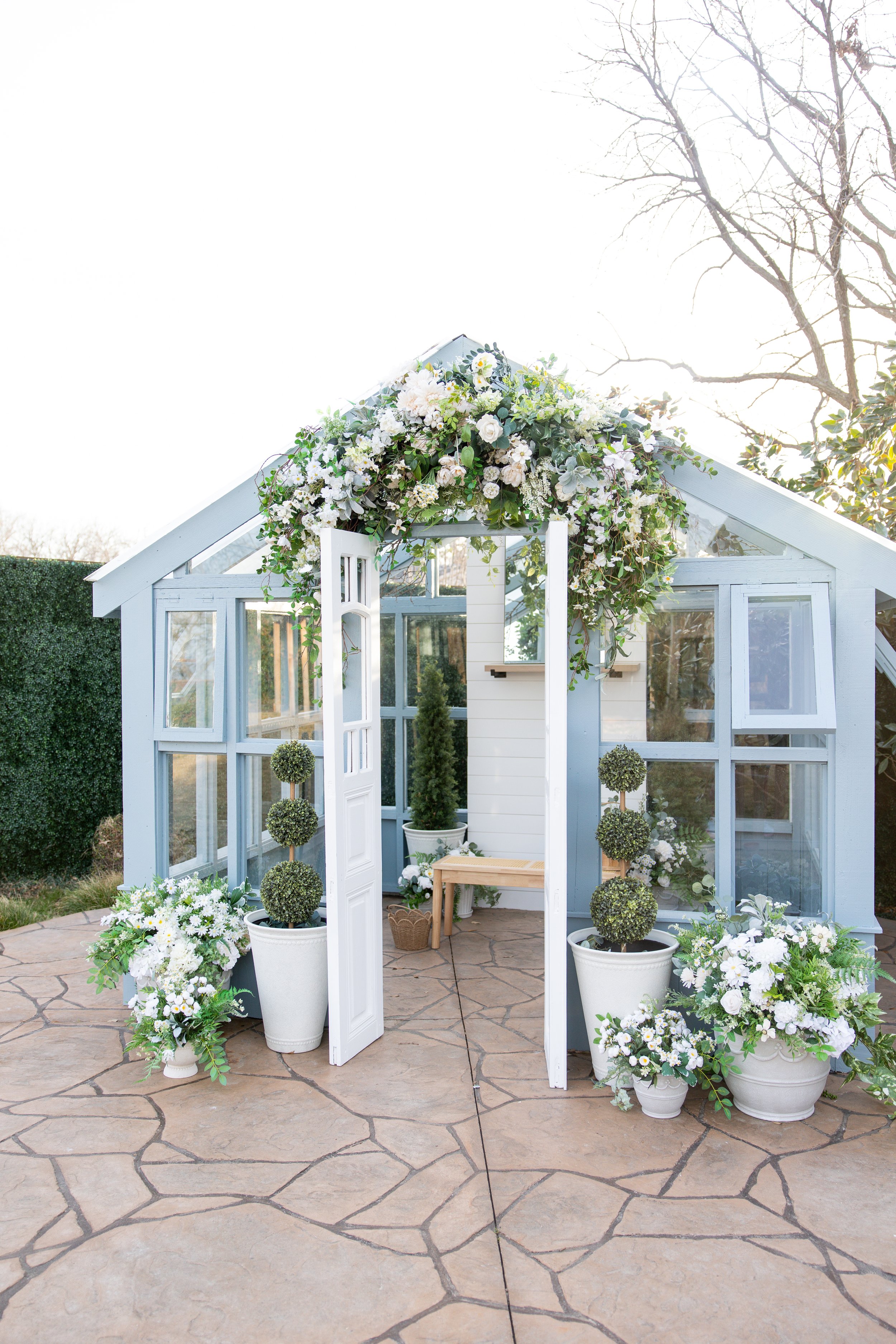 A small light blue wooden greenhouse decorated with white and green floral arrangements, including an arch of flowers above the entrance and potted greenery on either side, placed on a stone patio.