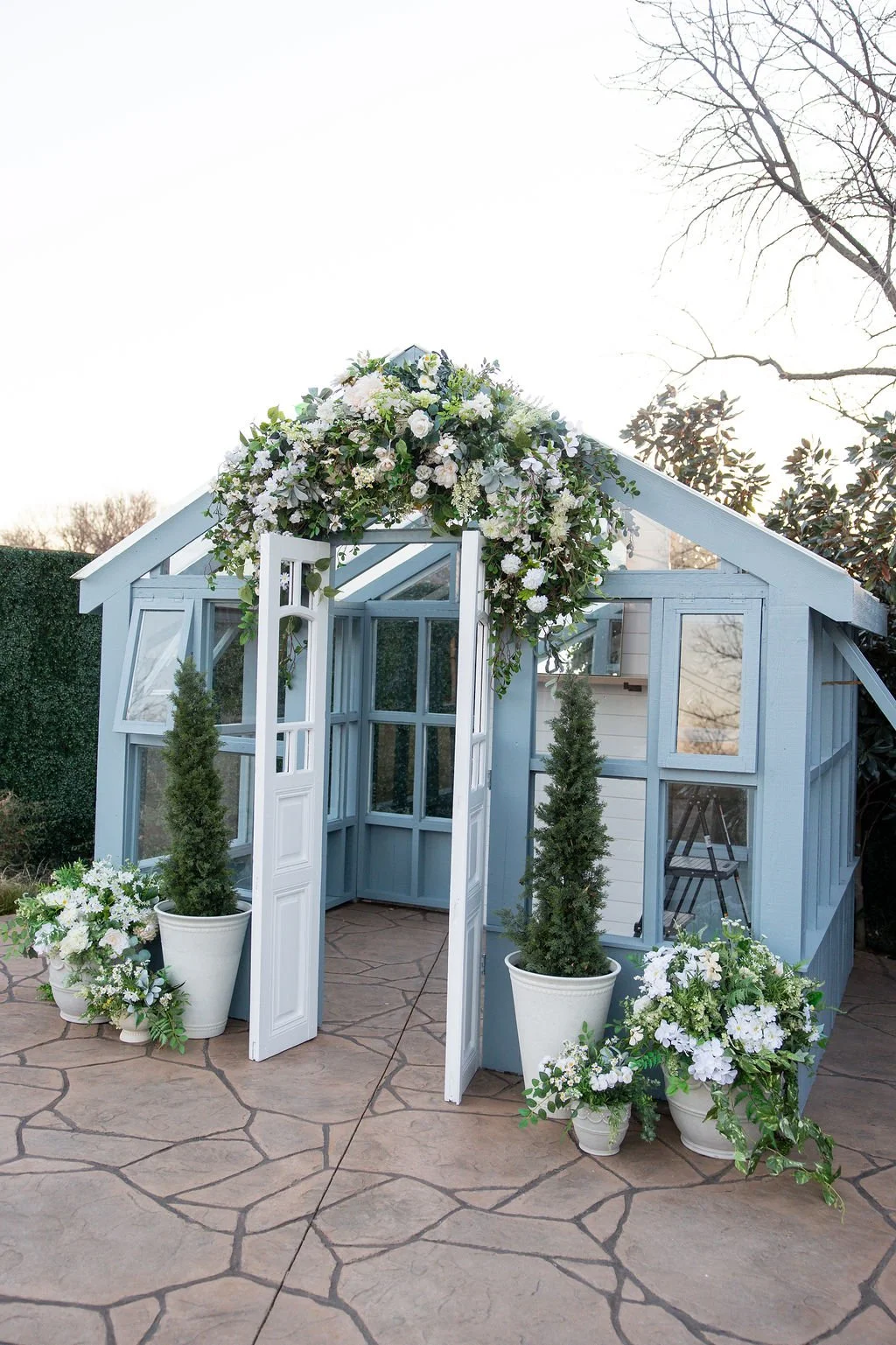 A small blue wooden greenhouse decorated with white flowers and greenery, including potted plants and floral arrangements, set on a paved patio.