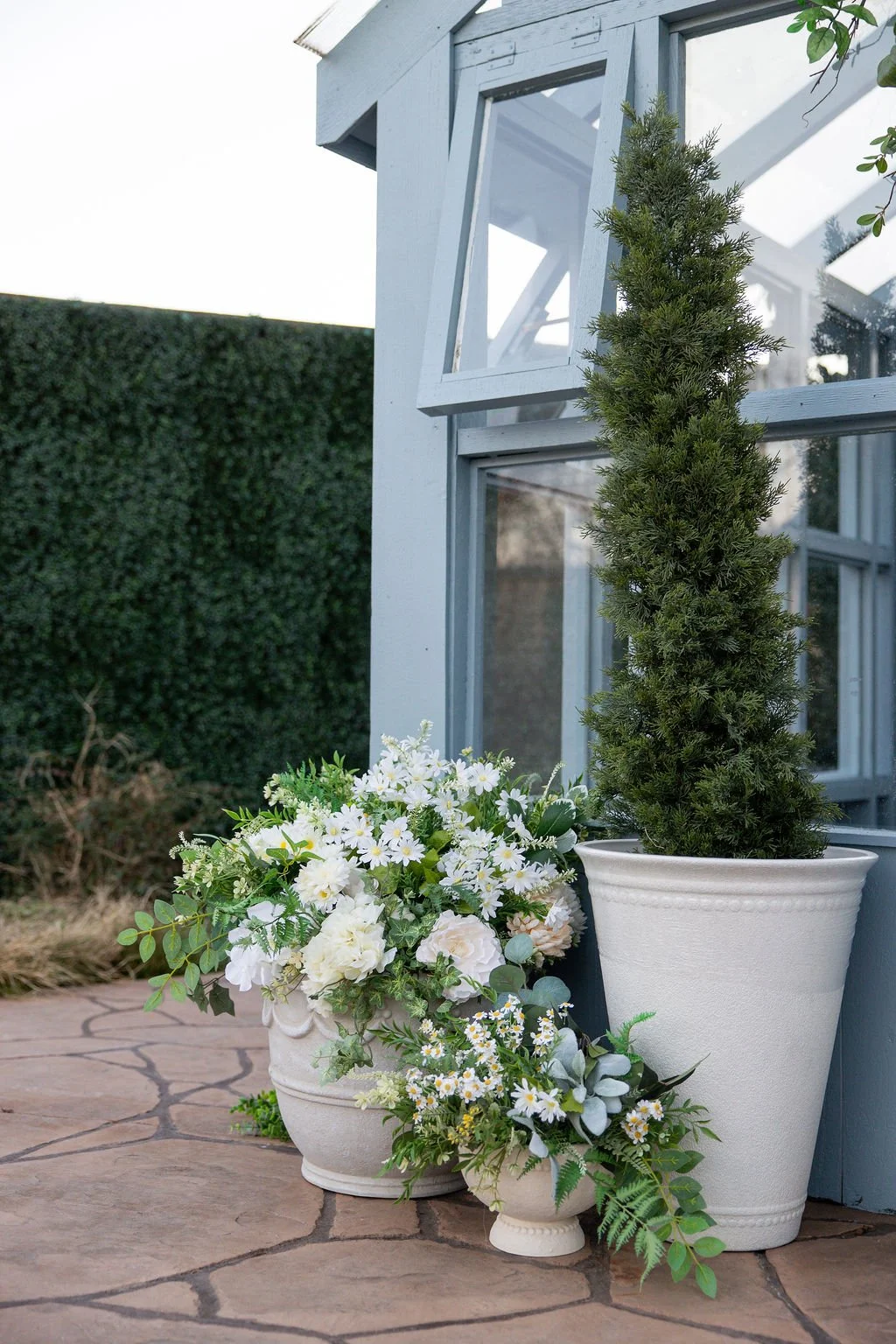 Potted evergreen tree and floral arrangement outside near a house with a glass window. The pots are white and decorative, placed on a stone patio.