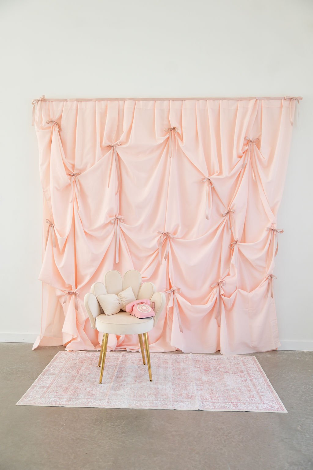 Pink fabric backdrop with tied bows and draping, beige chair with gold legs and pillows, pink rotary phone, all on a light pink rug on a gray floor.