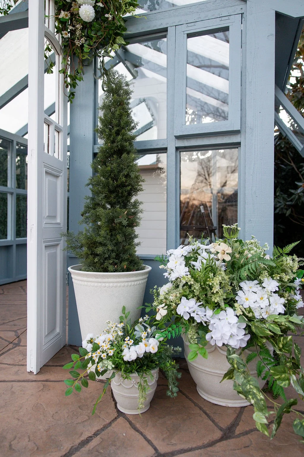 A small potted evergreen tree and white flowering plants in decorative pots arranged outside a light blue wooden greenhouse with glass panes and a white door.