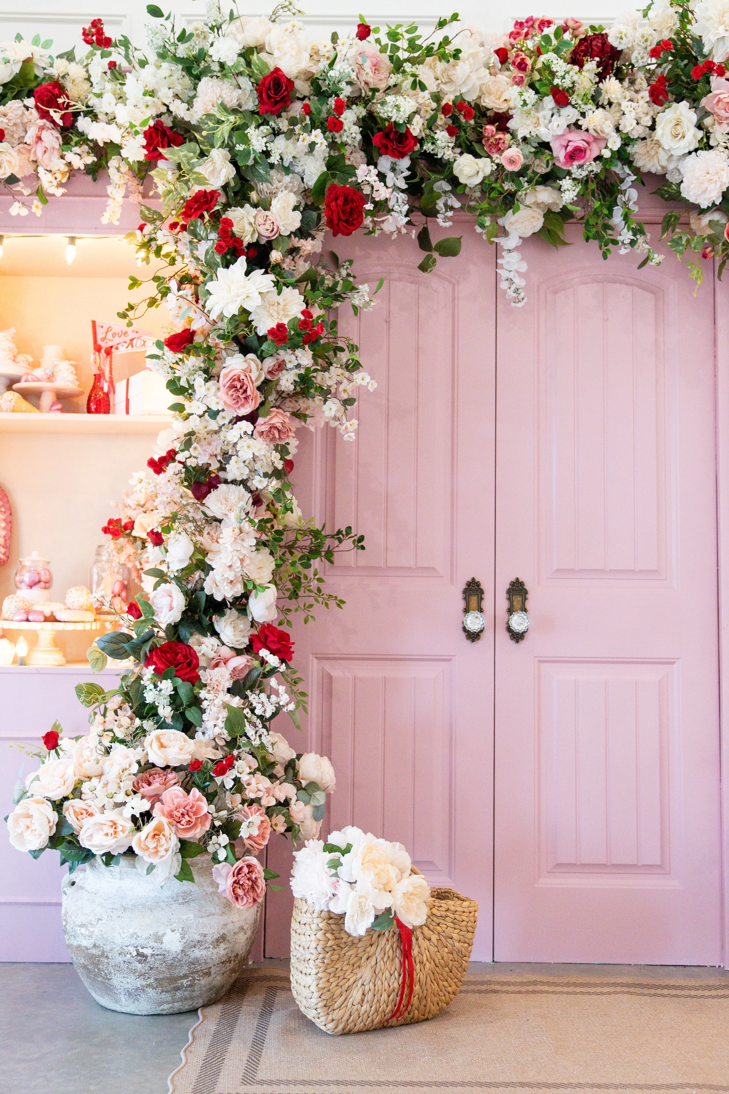 Pink double doors surrounded by a large floral arch with pink, red, white, and blush flowers, and a basket of white flowers on a woven bag on the floor.