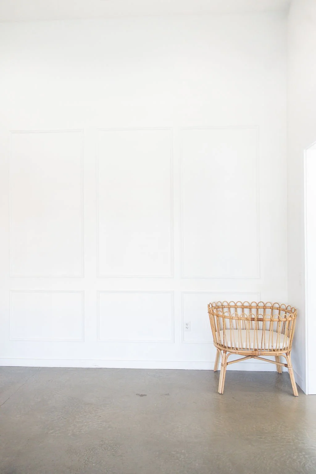 A corner of a room with white walls and a beige floor, featuring a wicker chair with a cushion placed against the wall.