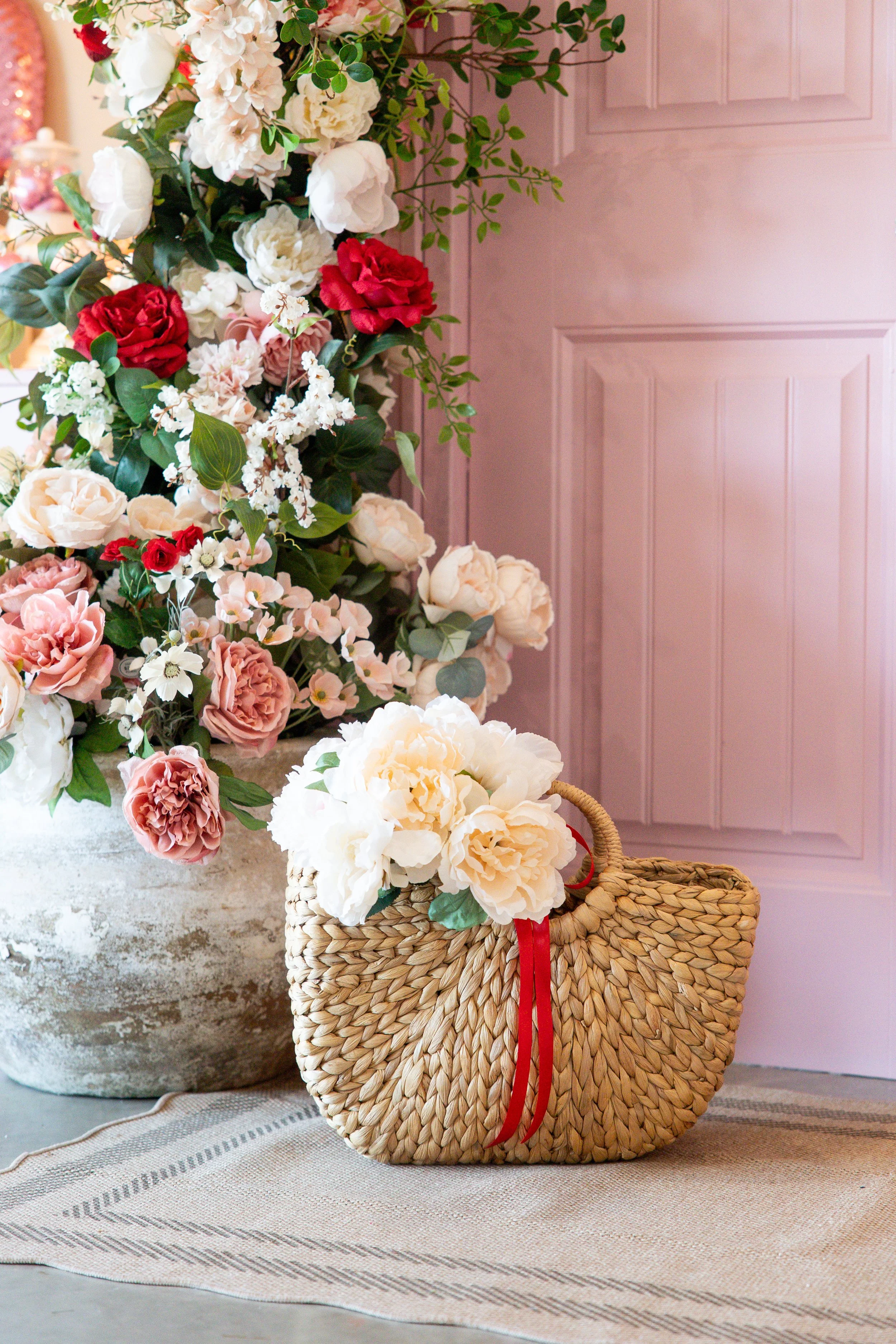 A large arrangement of pink, white, and red flowers in a rustic vessel and a woven straw handbag decorated with white peonies and greenery, placed on a striped cloth in front of a pink paneled door.