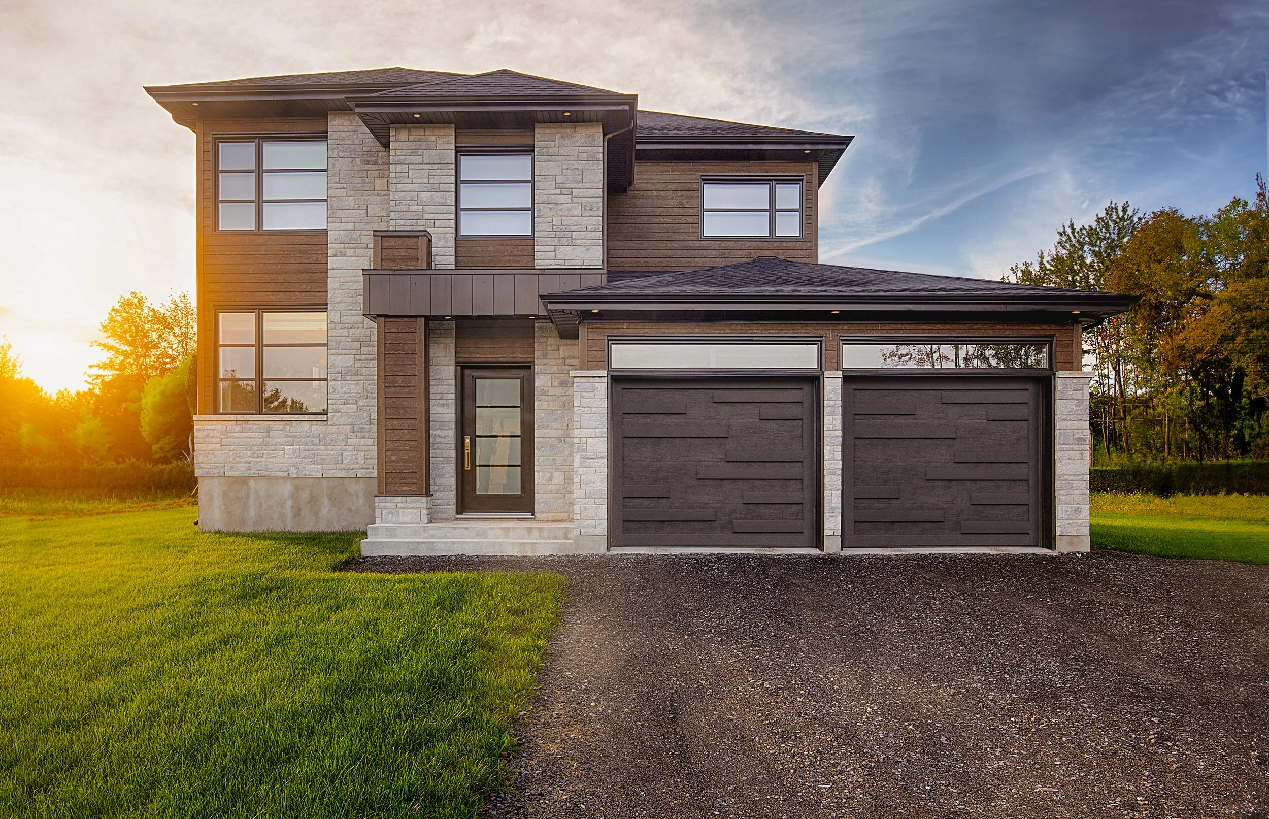 Modern two-story house with brick and wood siding, large windows, and a double garage, set against a sunset sky and surrounded by green grass and trees.