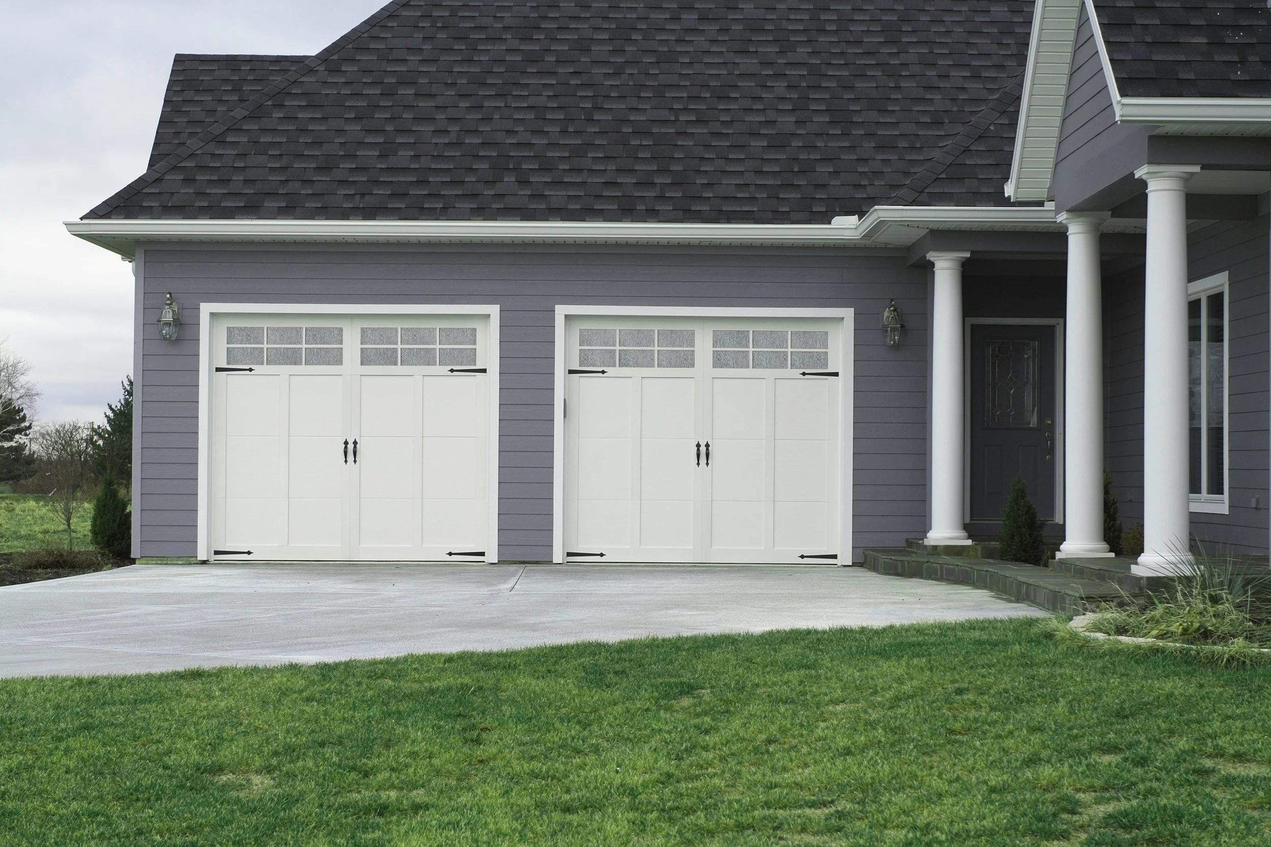 Front view of a house with a grey exterior, white garage doors, a small porch with white columns, and a well-maintained green lawn.