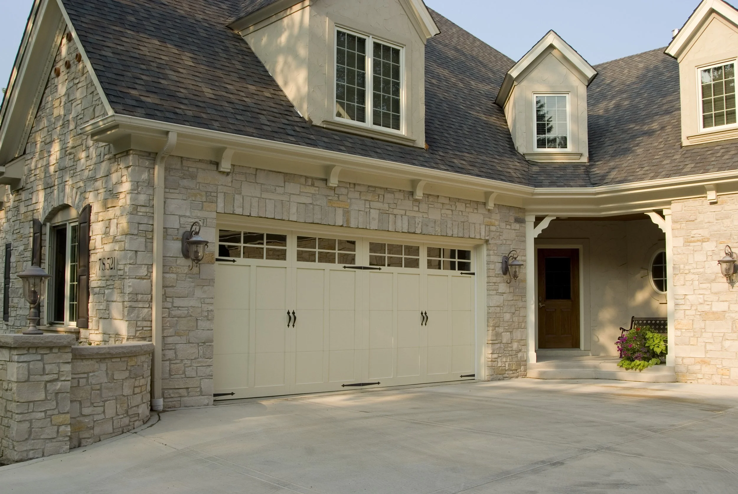 Front view of a house with a stone facade, a white garage door, and a small porch with a wooden door, an armchair, and potted plants in a sunny setting.