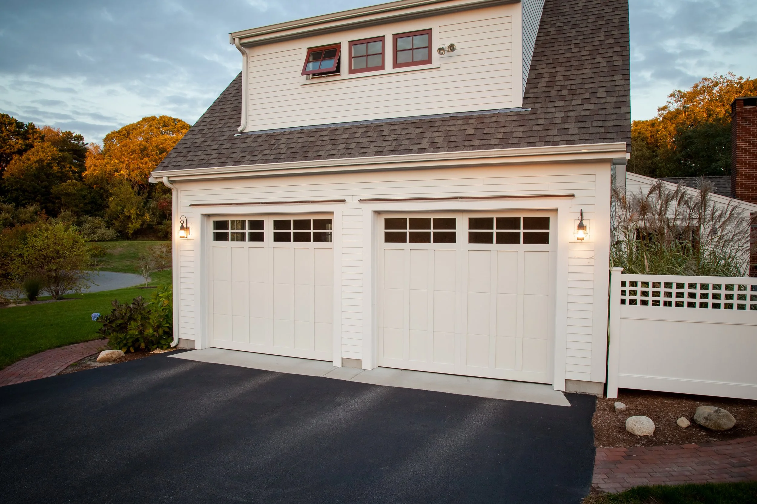 White double garage doors on a house with exterior lights and a paved driveway, surrounded by trees and shrubs during dusk.