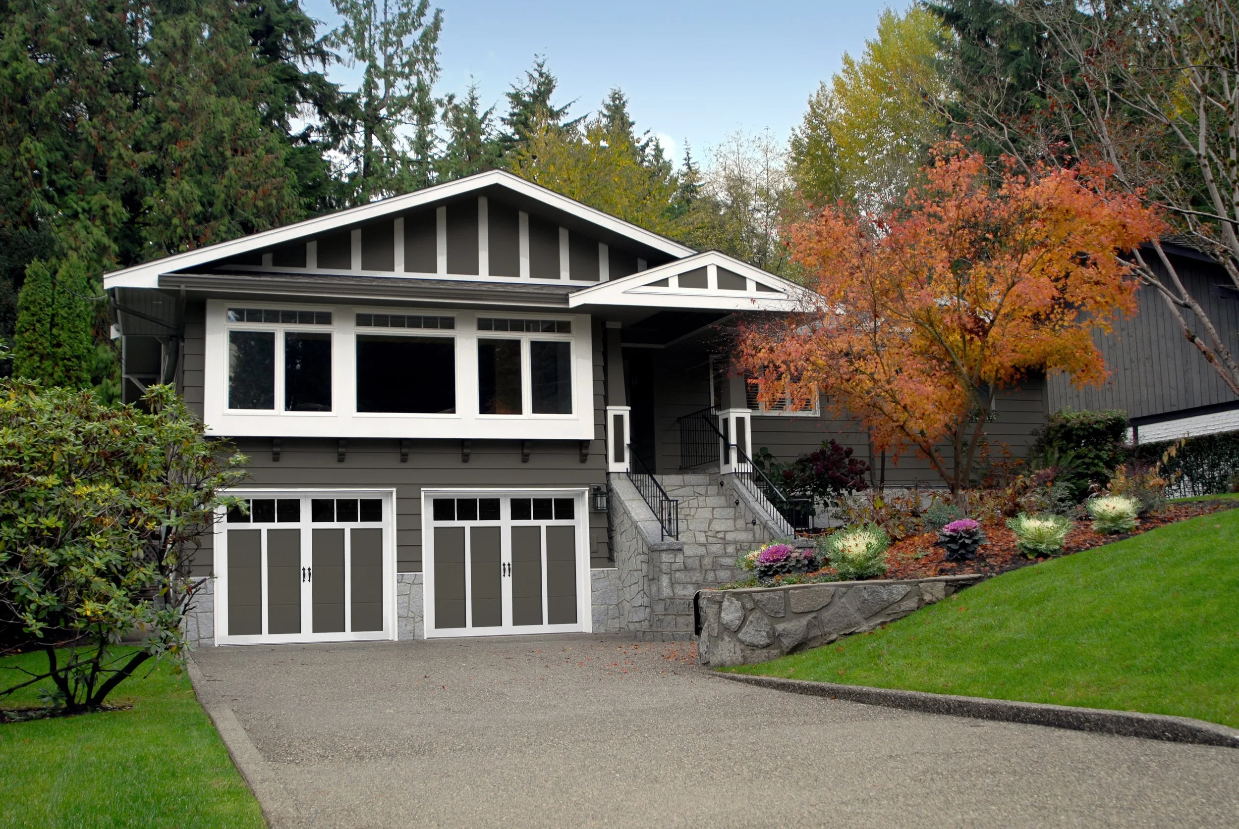 A two-story house with a gray exterior, white trim, and a front garden with colorful plants and a small tree with orange leaves, situated on a sloped lawn with a concrete driveway.