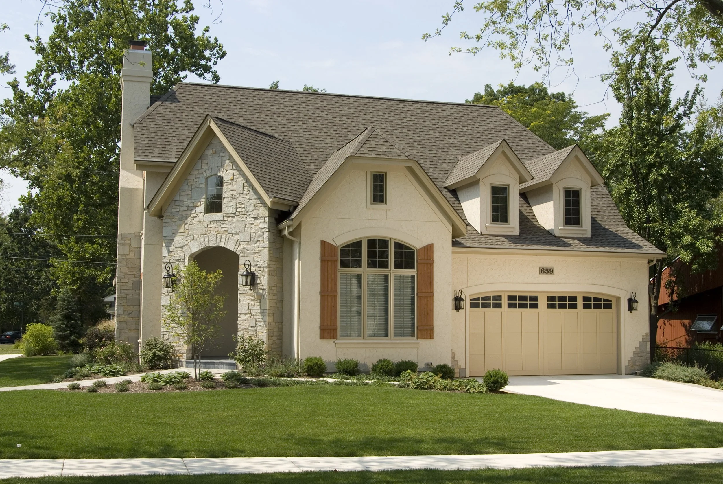 Front view of a two-story house with a stone and stucco exterior, brown shingled roof, large arched window with wooden shutters, and a garage with a raised panel door, surrounded by a well-maintained lawn and trees.