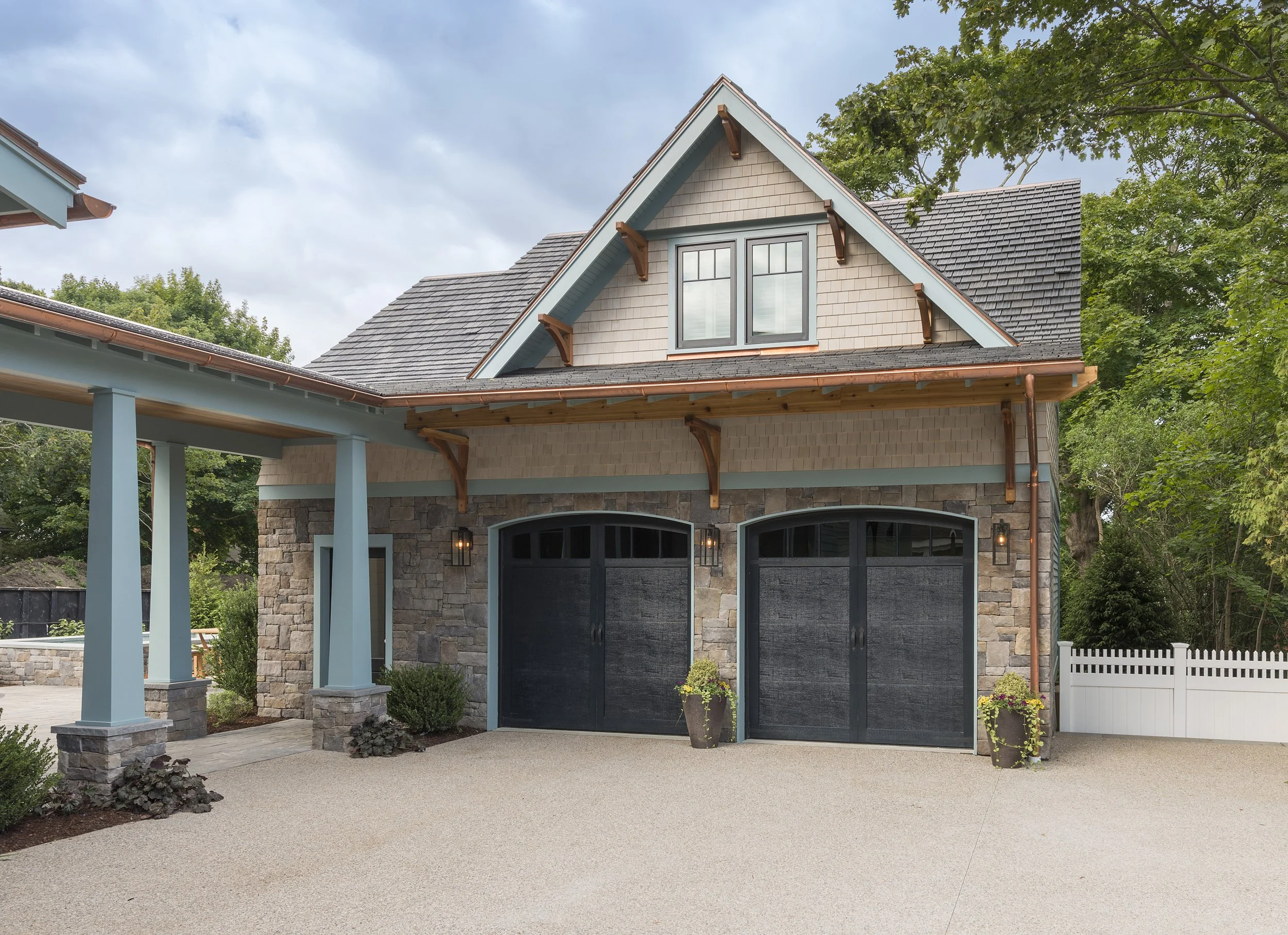 A modern house with a stone and pastel siding exterior, black garage doors, and a covered porch area with blue support beams and decorative wood braces, surrounded by greenery.