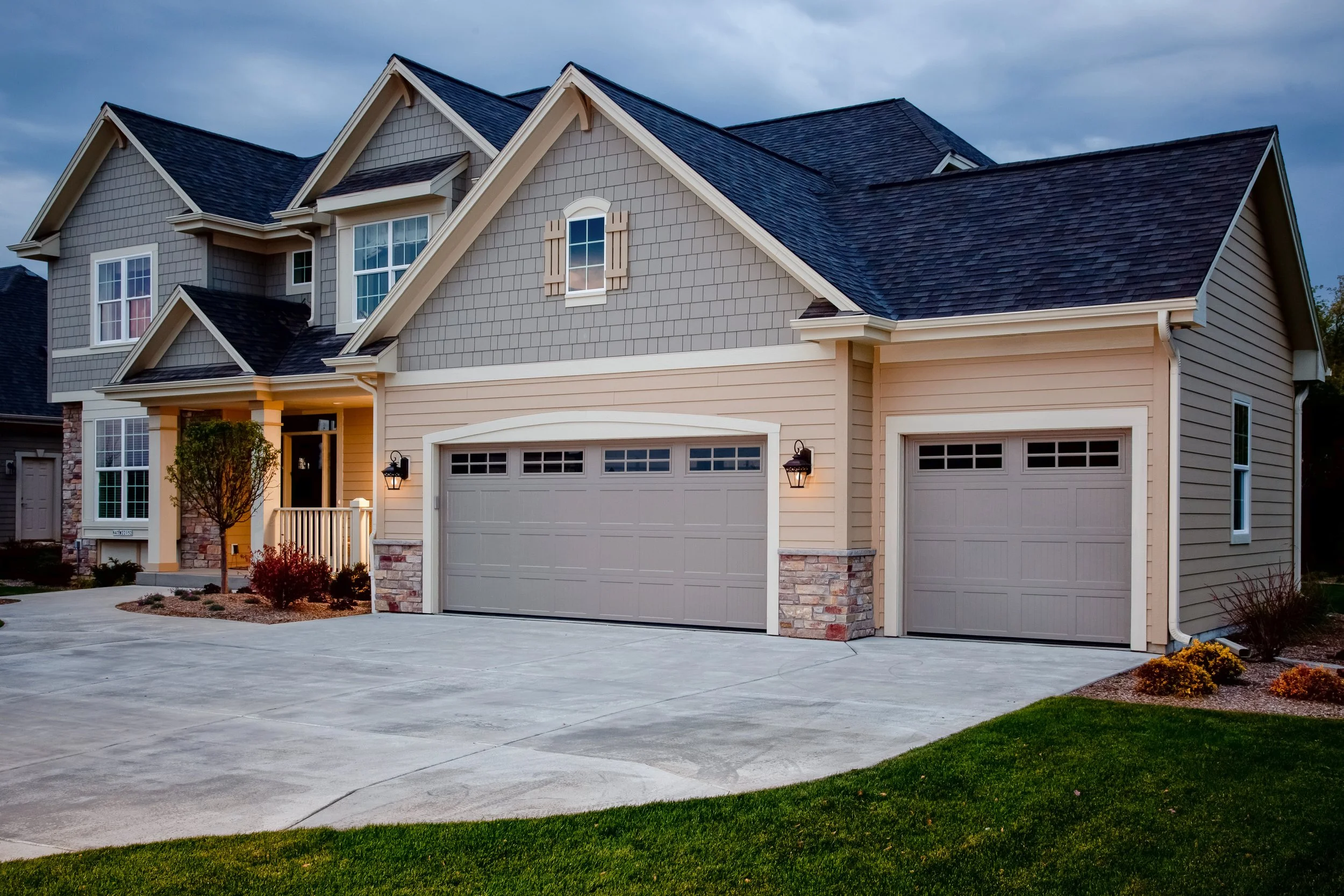 Front view of a two-story house with beige siding, gray garage doors, stone accents, and a well-manicured lawn. The house has multiple gabled roofs and a small porch with a tree and shrubs nearby.