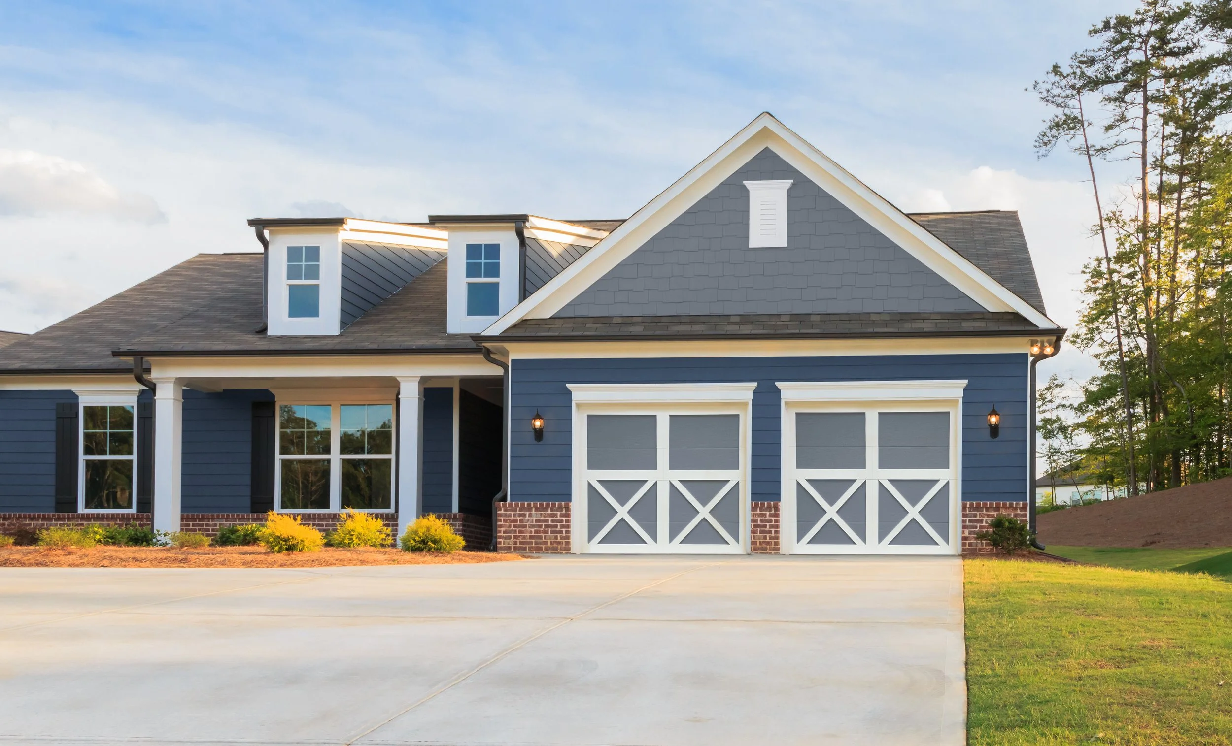 A modern two-story house with dark blue siding, white trim, a gabled roof, and a two-car garage, set against a partly cloudy sky and surrounded by greenery.