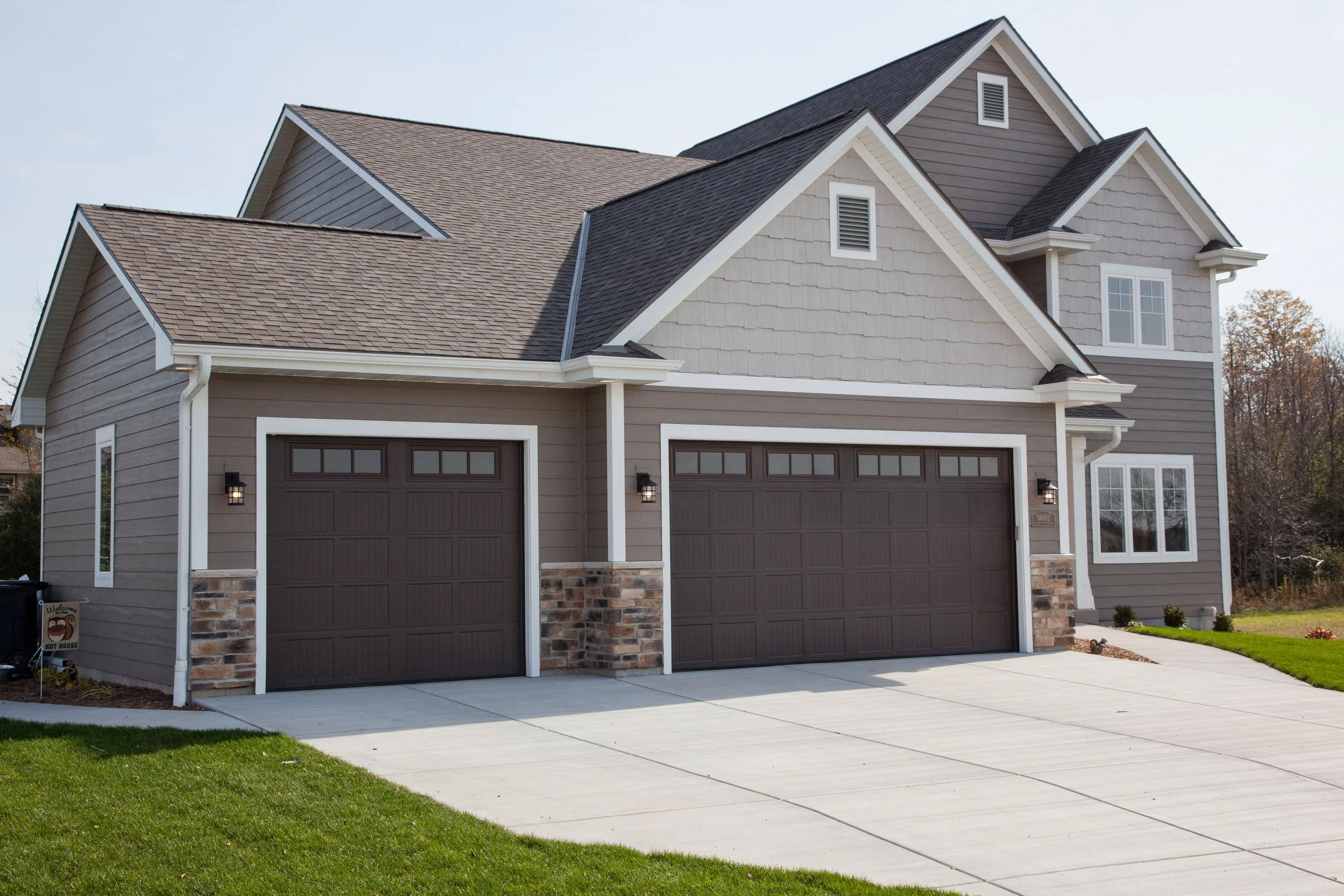 A modern two-story house with grey and tan siding, two attached garages having dark brown doors, and a concrete driveway. The house features several windows, gabled roofs, and a well-kept lawn.