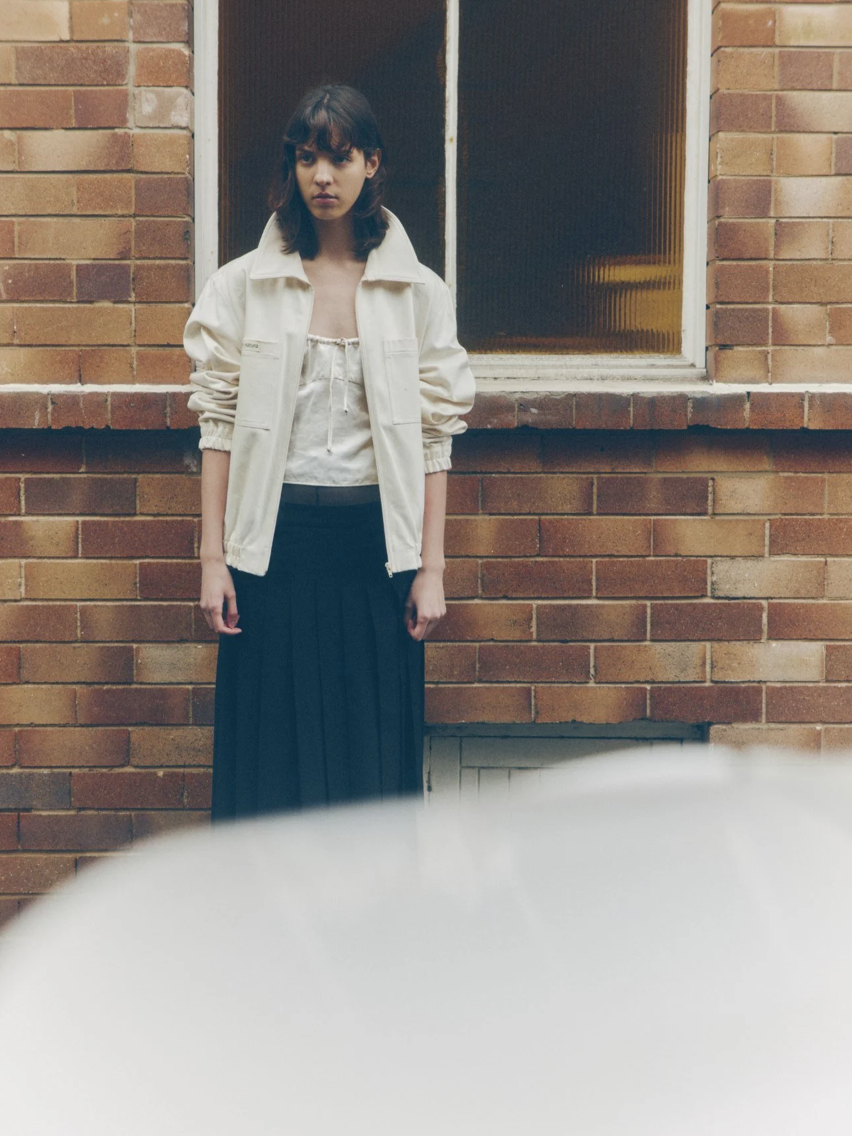 A young woman standing against a brick wall near a window, looking downward with a serious expression, wearing a beige jacket over a white top and a long black skirt.