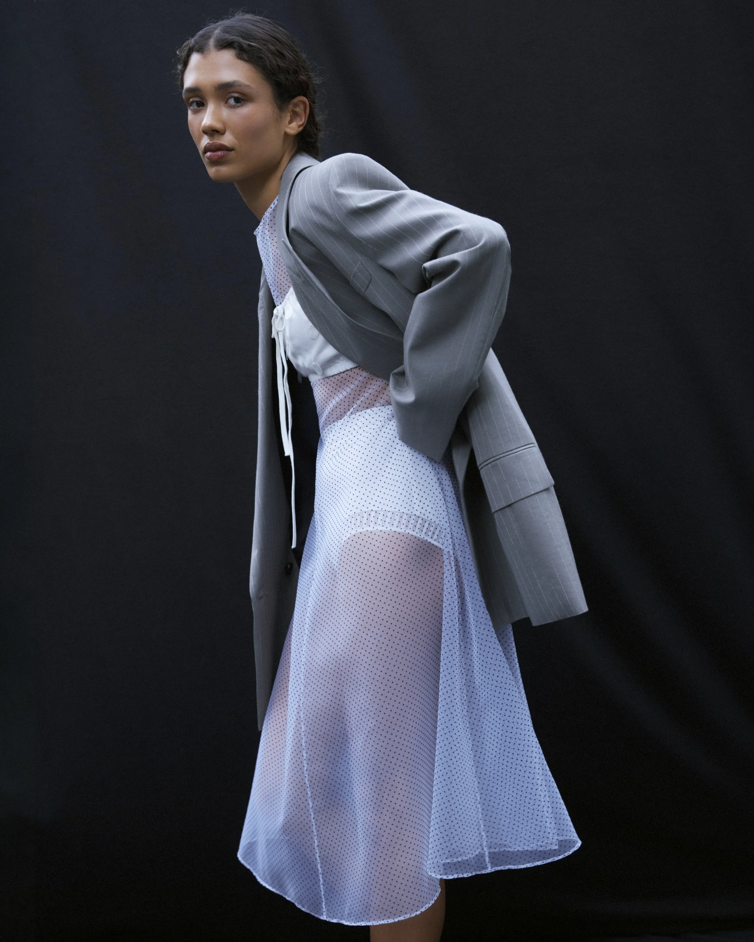 A woman wearing a gray pinstripe blazer over a white sheer dress with polka dots, standing against a black background.