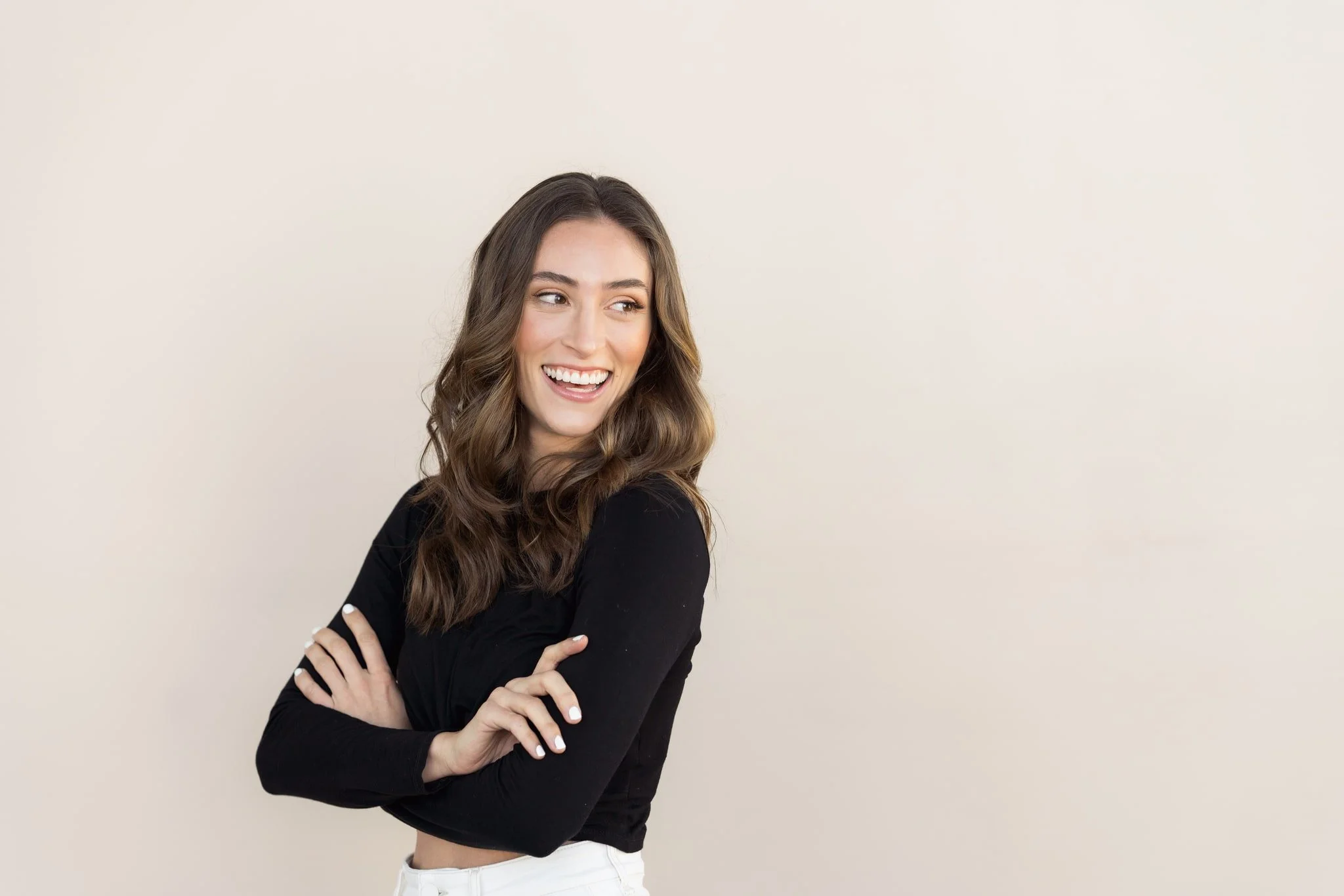 Shanon Casey, website designer, with long wavy brown hair, smiling, wearing a black long-sleeve top and white pants, standing against an off-white background.