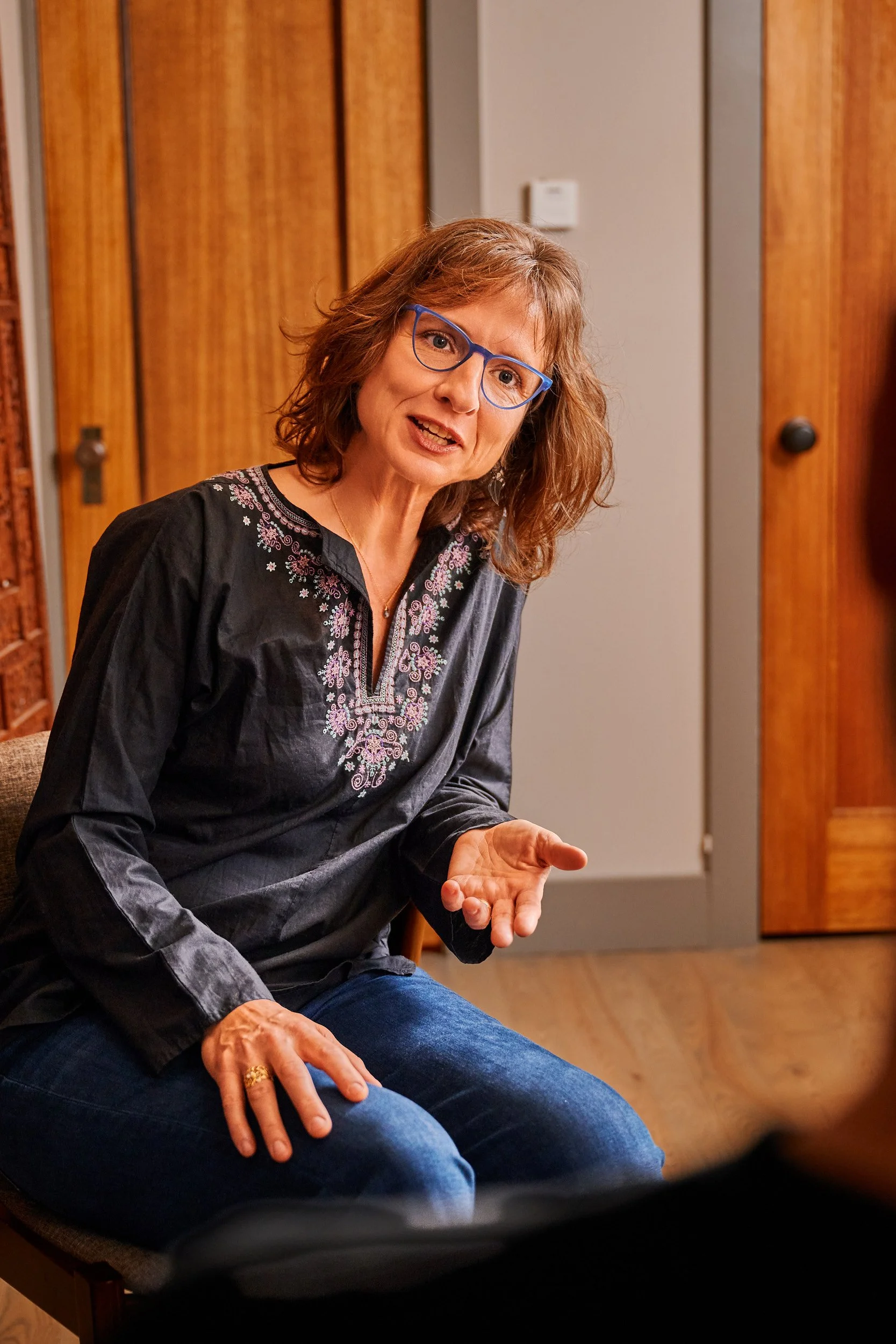 A middle-aged woman with curly brown hair, blue glasses, and a black embroidered blouse, sitting on a chair in a room with wooden cabinets, engaged in conversation or giving a presentation.