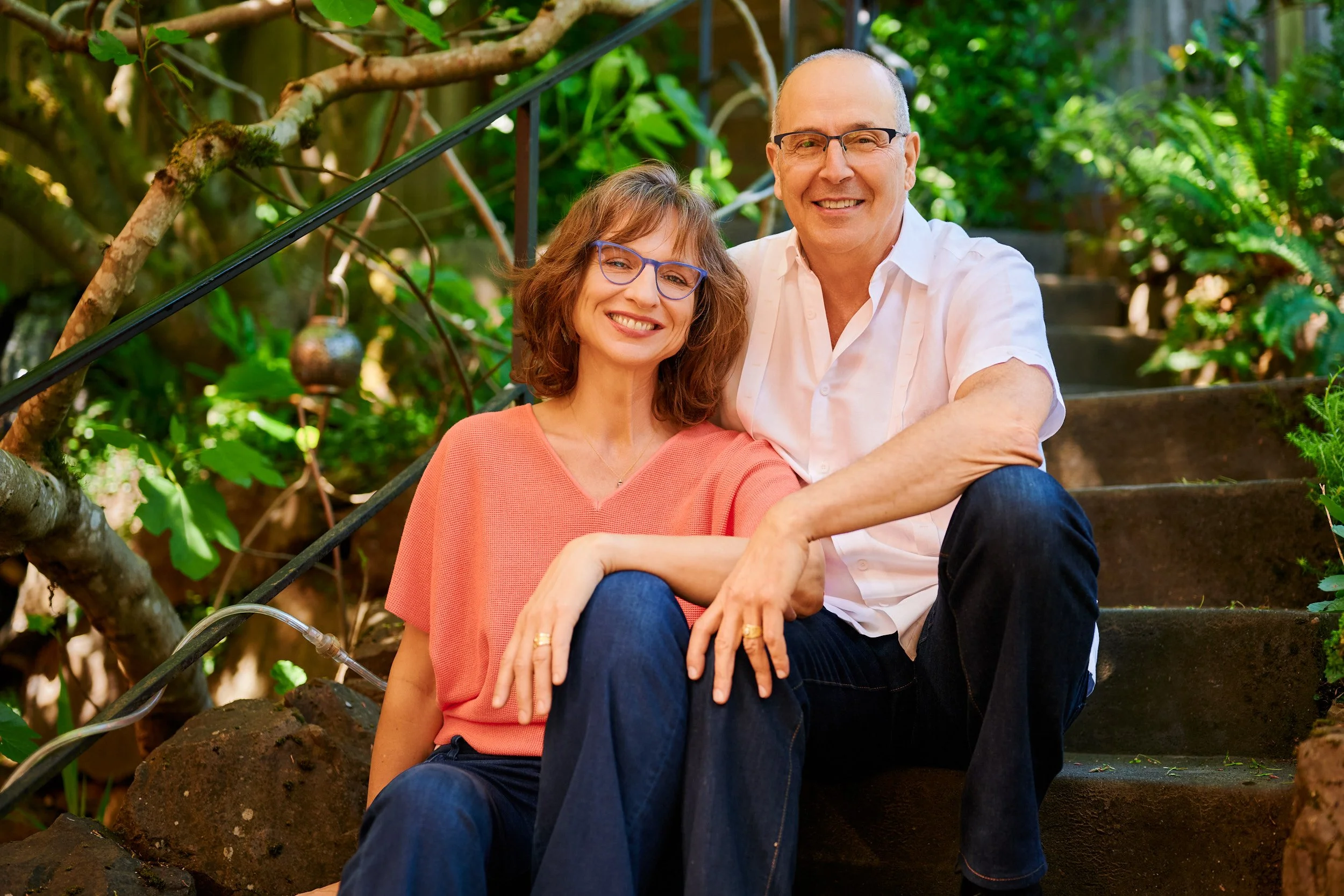 A smiling middle-aged couple sitting on outdoor steps surrounded by green plants and trees, wearing casual clothes and glasses.