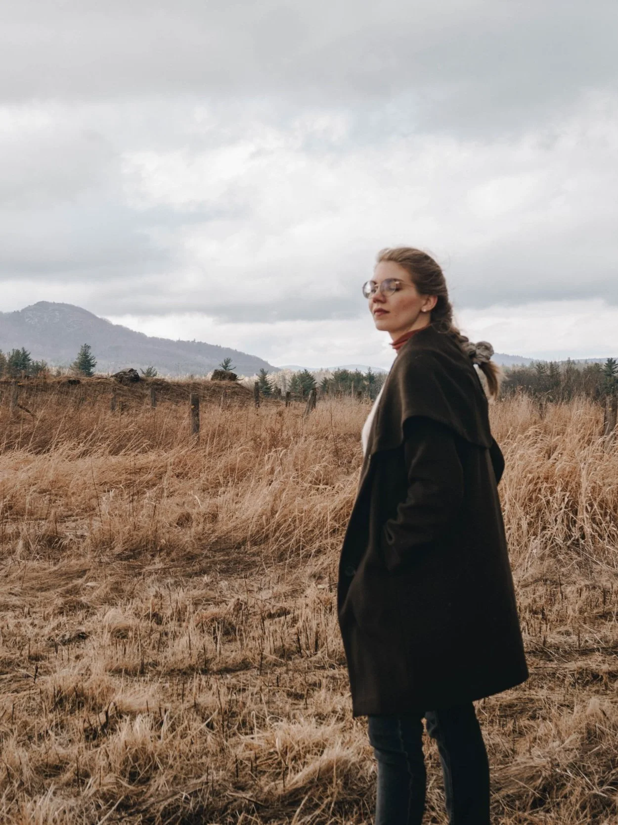 A woman with long blonde hair in a braid, wears a dark green coat. Behind her is a wheat colored field and in the background there are mountains.