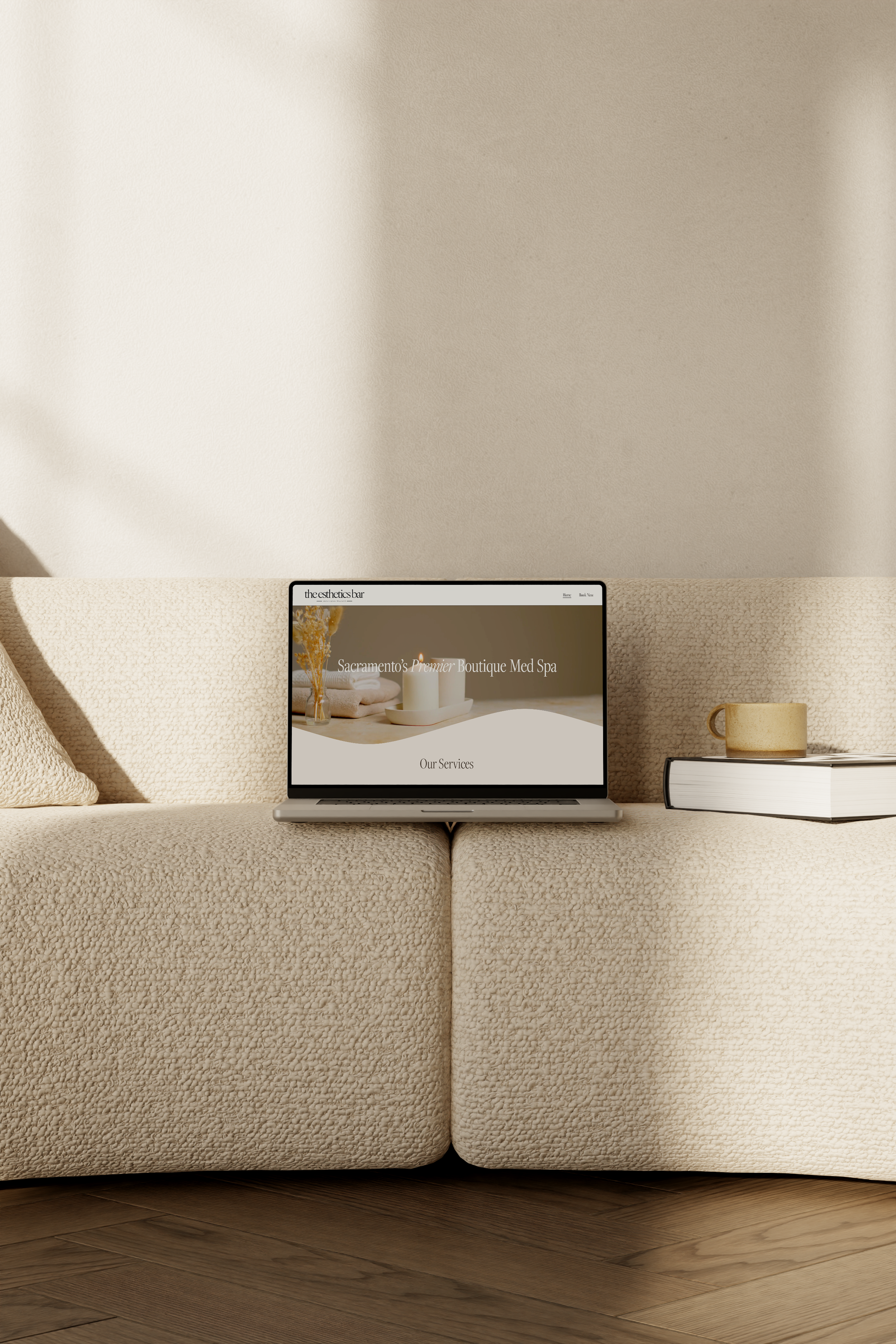 Couch with beige textured fabric in a living room, with a laptop on the center cushion displaying a spa website, a golden mug on the right, and two stacked books underneath it. The background is a beige wall with shadow on the left side.