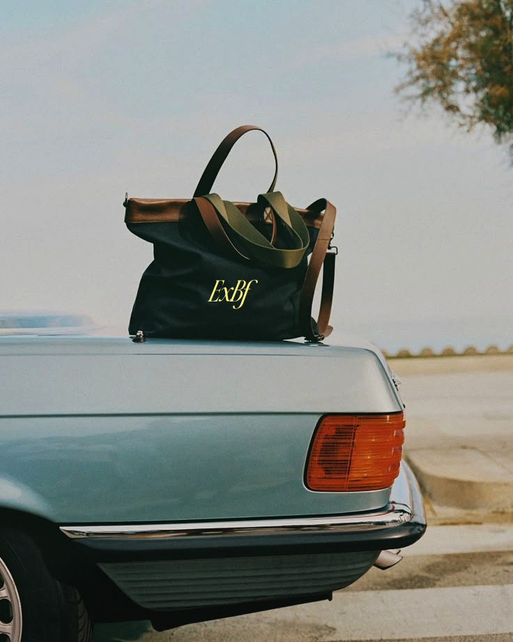 A black tote bag with green straps and brown handles, featuring the yellow text 'ExbF', sits on the trunk of a vintage silver car in an outdoor parking lot.