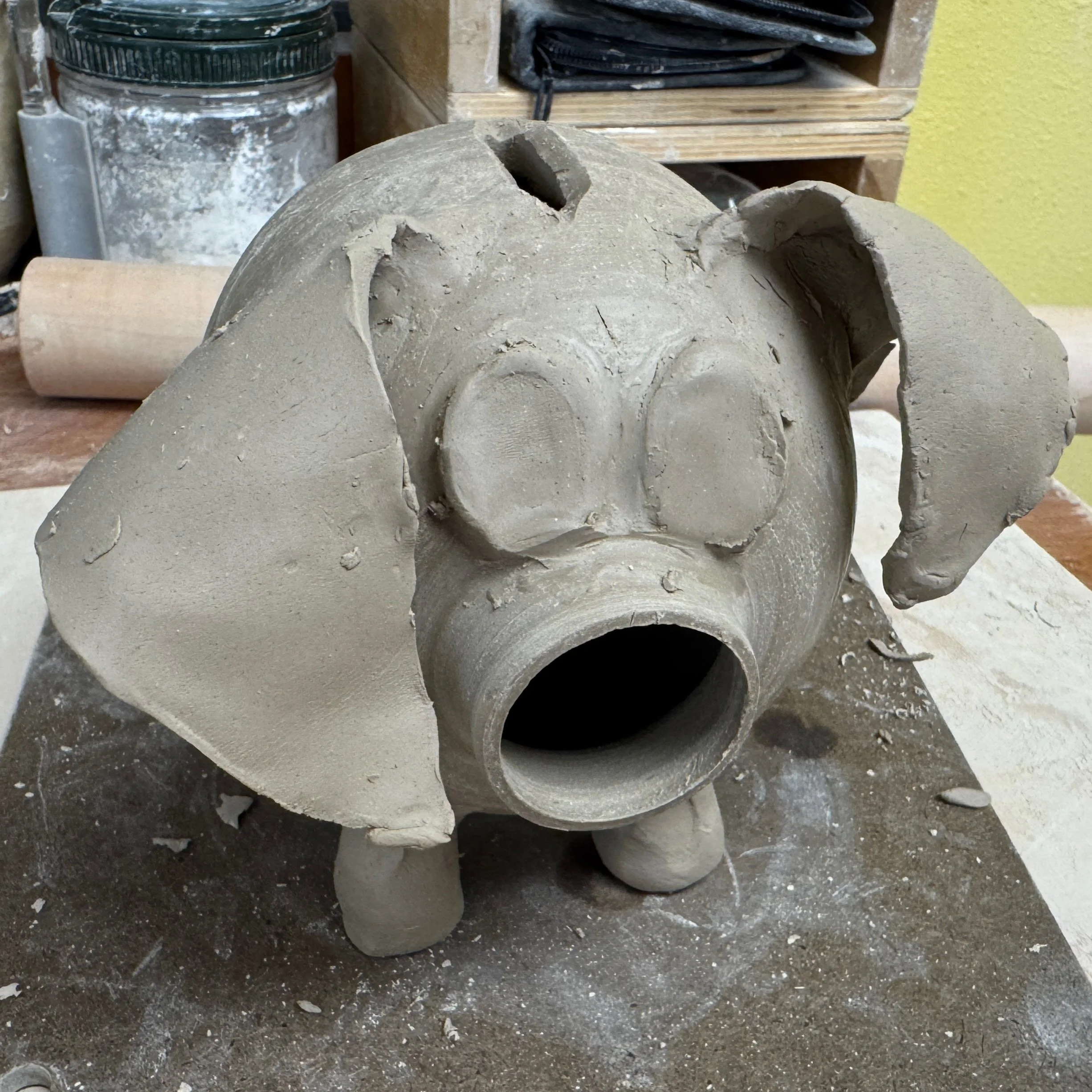 A clay piggy bank in progress, featuring a wide, open hole as a nose, big flat eyes, and floppy ears.