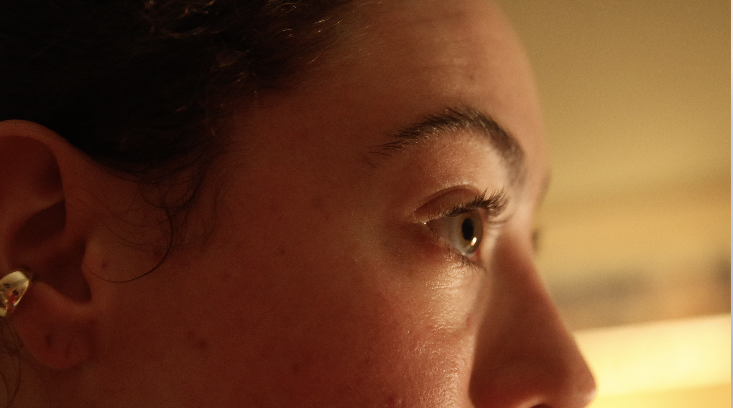 Close-up side profile of a woman's face showing her eye, eyebrow, and part of her ear, with warm lighting.