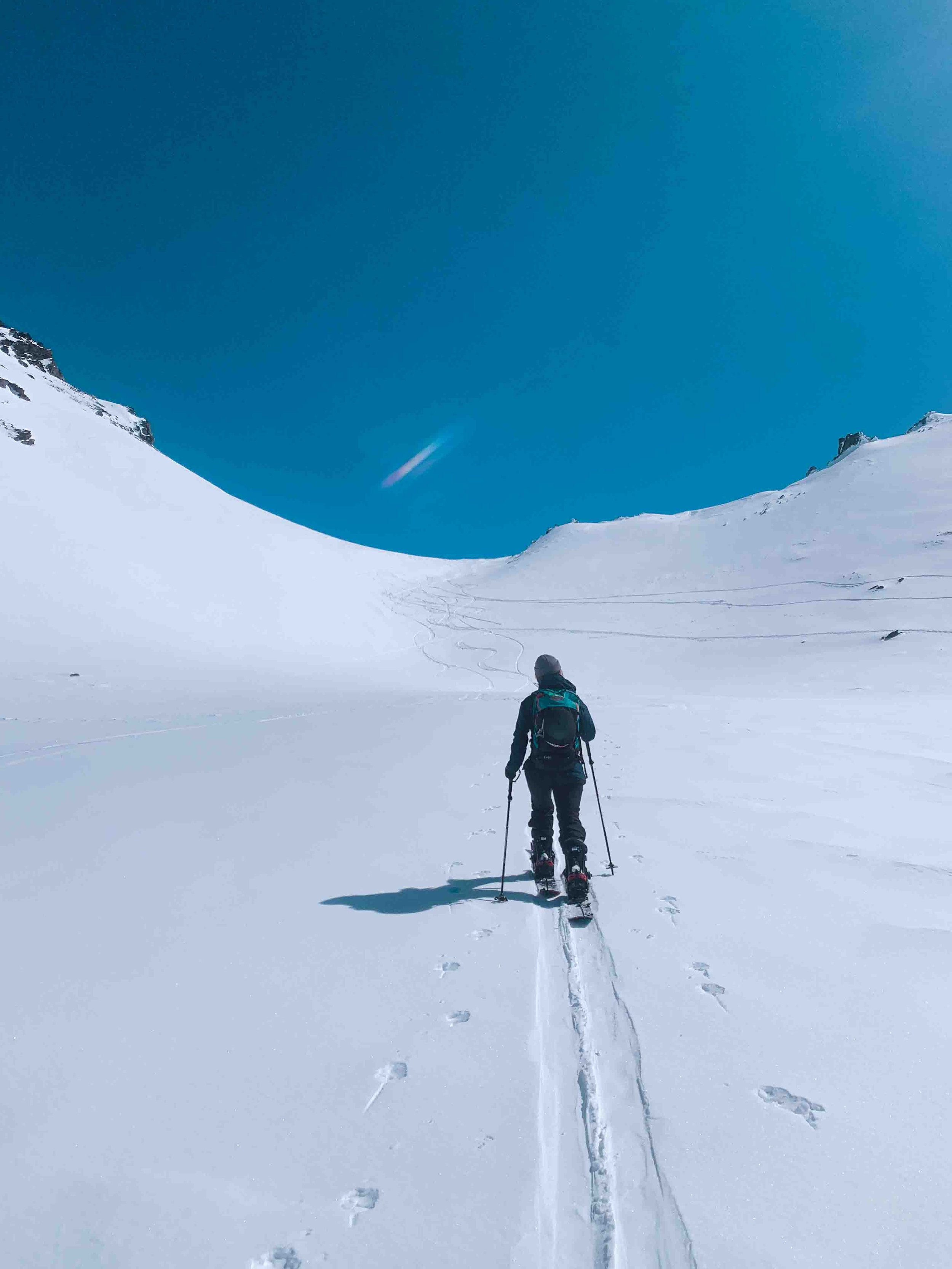 Person in winter clothing and ski boots skiing across a snow-covered alpine landscape with mountains in the distance and a clear blue sky.