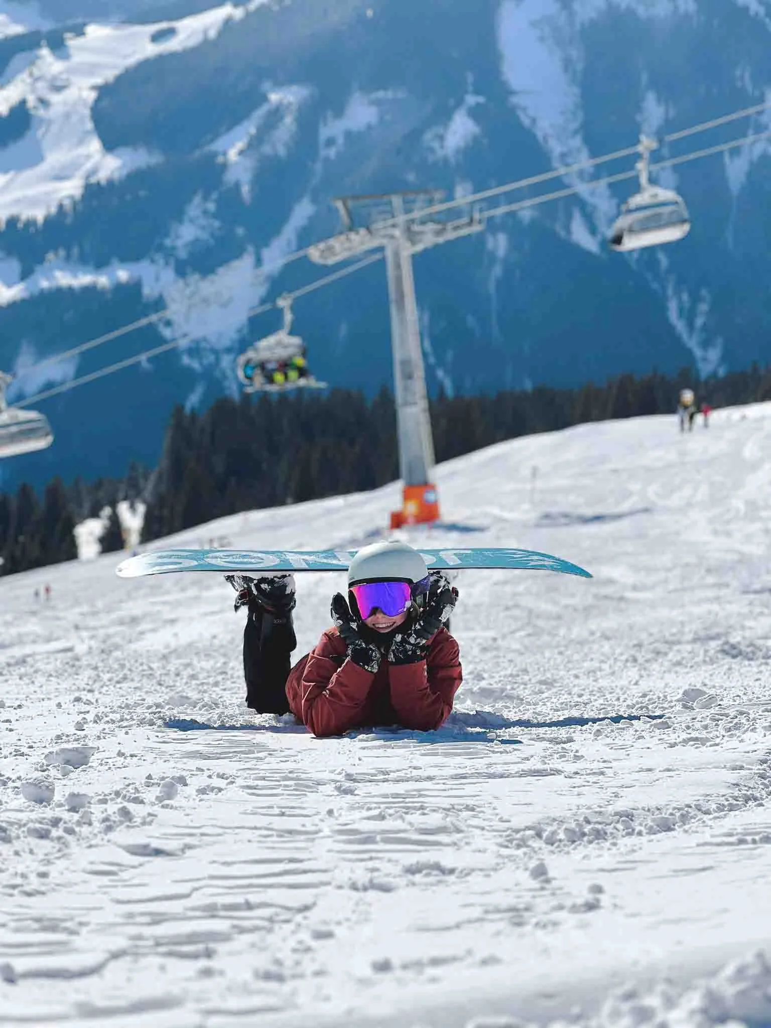 A person in snow gear lying stomach down  in the snow on a ski slope, mermaid pose with their snowboard attached to their feet with ski lifts and snow-covered mountains in the background.